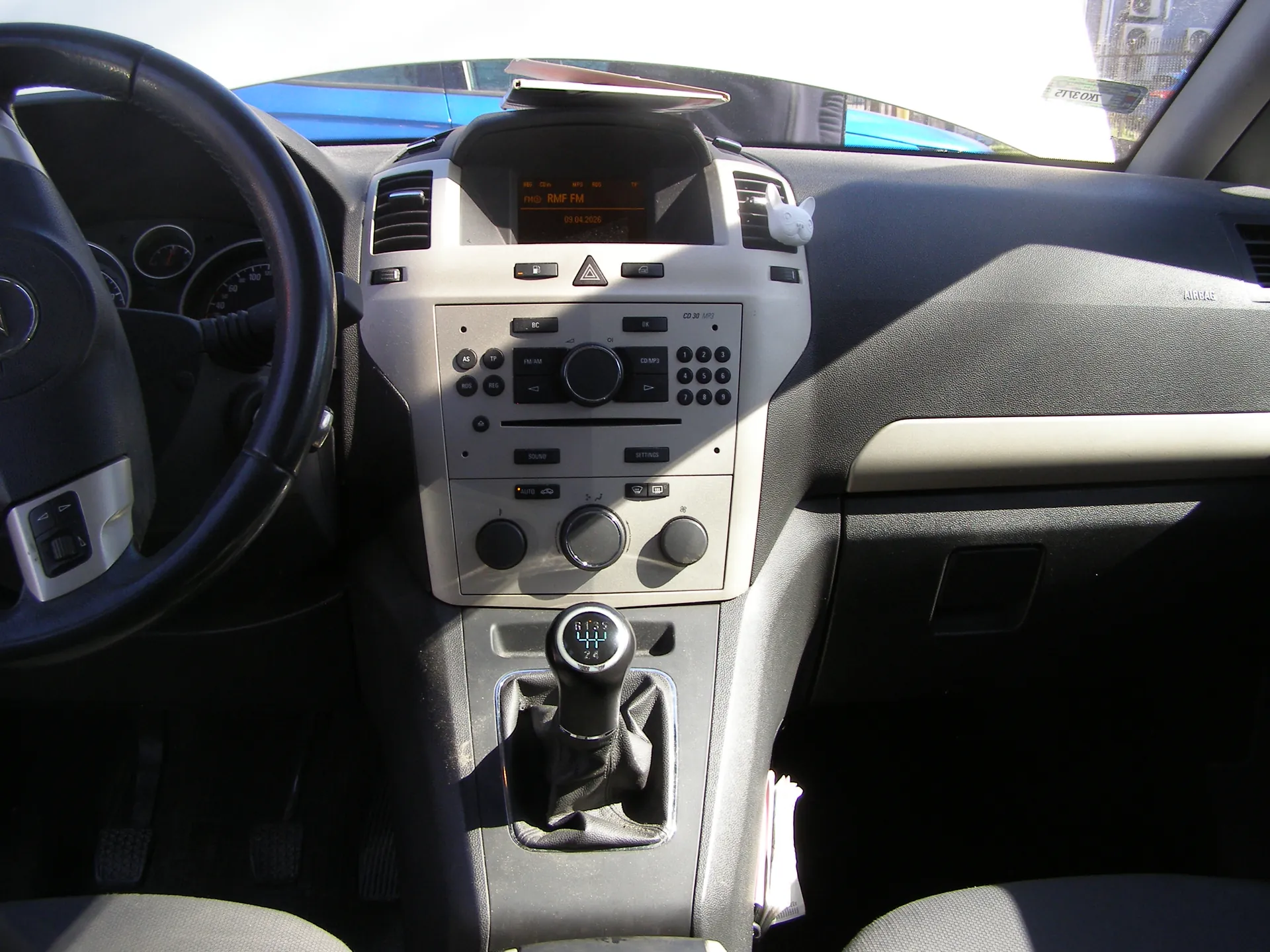 Wnętrze samochodu z widokiem na kierownicę po lewej stronie, deski rozdzielczej i centralnej konsoli. Na środku konsoli znajduje się radio z wyświetlaczem pokazującym 'RMF FM' oraz datą '09.04.2006', przyciskami sterującymi i suwakami. Poniżej radio widoczna jest automatyczna skrzynia biegów z wyświetlaczem 'P R N D 3 2 1'. Na desce rozdzielczej umieszczono białą figurkę zwierzęcia. W tle przez przednią szybę widać niebieski samochód i fragment budynku. Siedzenia są szare, a wnętrze dominują odcienie czerni i szarości.