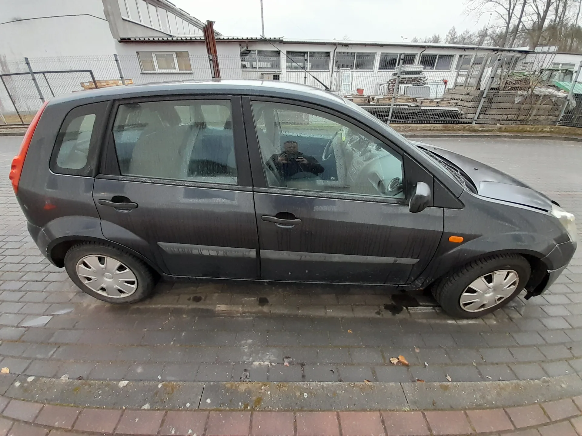 Ciemnoszary samochód osobowy typu hatchback zaparkowany na brukowanej powierzchni. Samochód jest zabrudzony, z widocznymi smugami na karoserii. Przednia prawa fura jest uszkodzona, a maska otwarta. Wewnątrz pojazdu, przez przednią szybę, widać osobę trzymającą urządzenie do robienia zdjęć. W tle budynek z białymi ścianami i oknami, ogrodzenie metalowe oraz stosy materiałów budowlanych. Na ziemi rozrzucone są suche liście, a niebo jest pochmurne.
