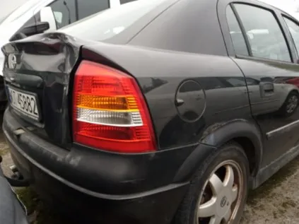 Czarny samochód osobowy typu sedan widziany od tyłu z widocznym uszkodzeniem bagażnika. Na tablicy rejestracyjnej widoczny napis M 4598Z. Czerwone i pomarańczowe światło tylnego reflektora, koło z felgą, podwozie na żwirowym podłożu. W tle częściowo widoczny inny pojazd oraz budynek.