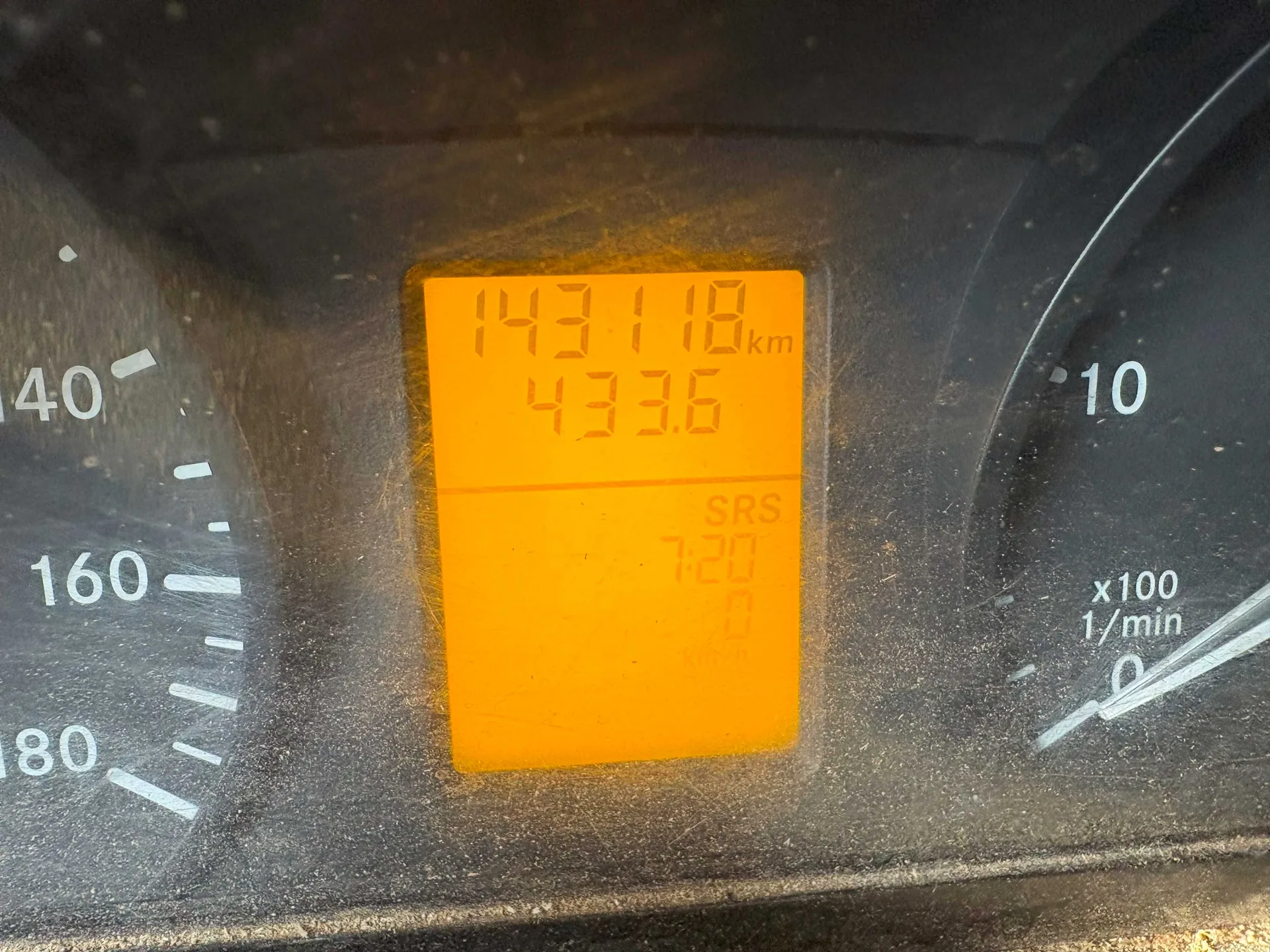Zbliżenie na deski rozdzielcze samochodu z pomarańczowym wyświetlaczem cyfrowym. Wyświetlacz pokazuje przebieg 143118 km, poniżej 433.6, napis SRS, godzinę 7:20 oraz wartość 0. Po lewej stronie widoczny wskaźnik prędkości z oznaczeniami 40, 160, 180, po prawej tachometr z oznaczeniami 10 i x100 1/min. Wyświetlacz i deska rozdzielcza są lekko zabrudzone.
