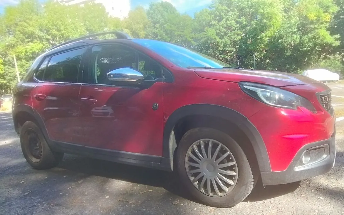 Czerwony samochód typu crossover zaparkowany na utwardzonym placu na tle zielonych drzew. Pojazd jest pokazany z profilu, z widocznymi srebrnymi lusterkami bocznymi, czarnymi plastikowymi nadkolami oraz wieloramiennymi felgami aluminiowymi.
