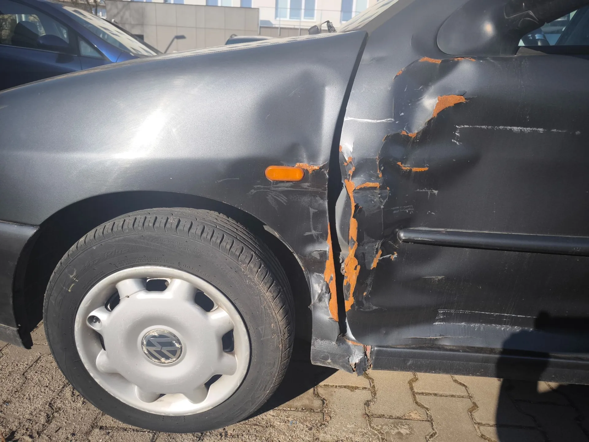 Zbliżenie na uszkodzony przód i drzwi kierowcy szarego samochodu marki Volkswagen. Widoczne są głębokie wgniecenia i zarysowania lakieru na drzwiach oraz błotniku, odsłaniające pomarańczową warstwę podkładu. Samochód ma srebrną felgę z logo Volkswagena i jest zaparkowany na kostce brukowej.
