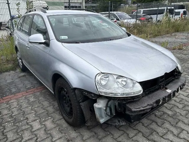 Srebrny samochód osobowy typu kombi zaparkowany na kostce brukowej. Pojazd jest uszkodzony z przodu – brakuje przedniego zderzaka, odsłaniając elementy konstrukcyjne i wzmocnienie. Samochód ma czarne felgi stalowe. W tle widoczne są inne zaparkowane samochody oraz ogrodzenie z siatki.
