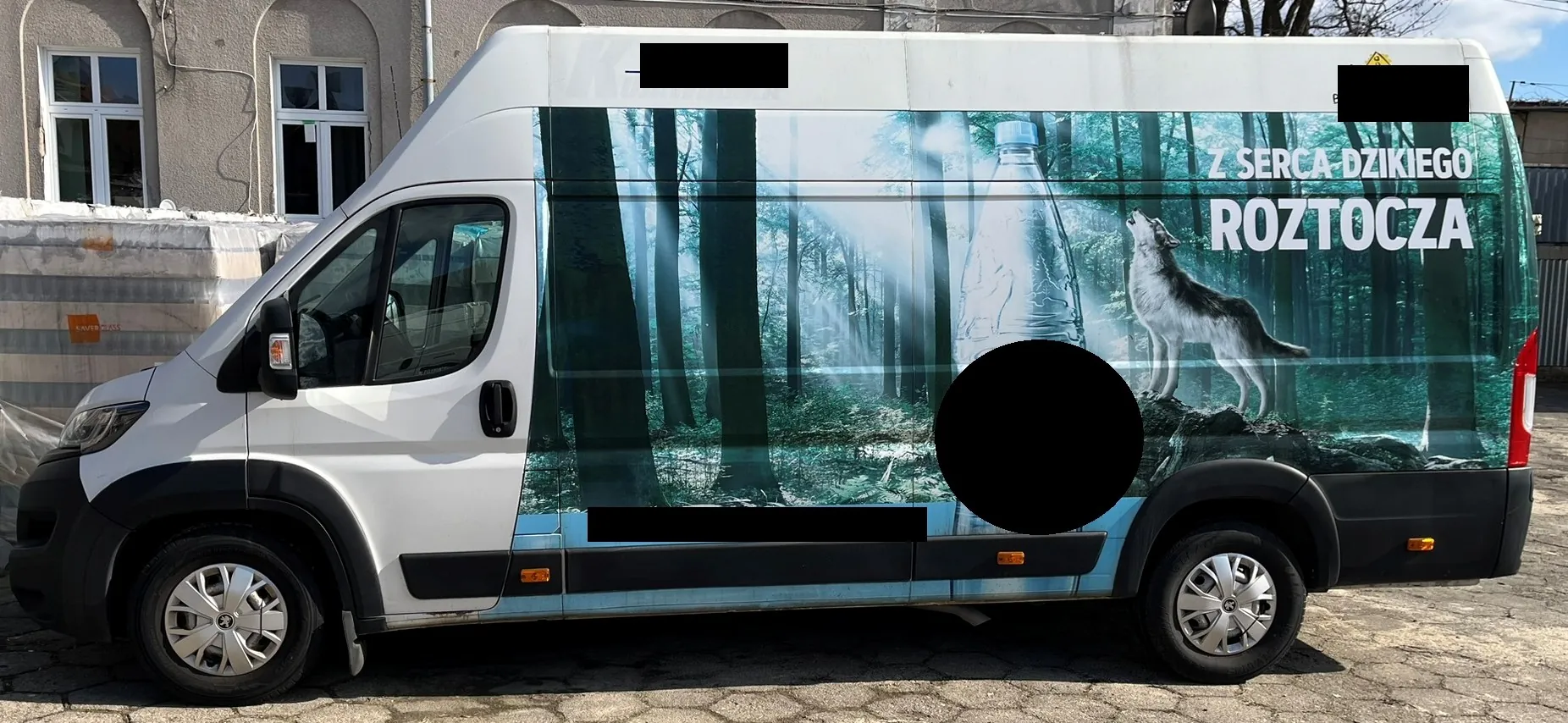 Biały samochód dostawczy z oklejeniem reklamowym na boku nadwozia. Grafika przedstawia gęsty, mglisty las z dużym wizerunkiem butelki wody oraz wilkiem stojącym na skale. Na obrazie widnieje napis: 'Z SERCA DZIKIEGO ROZTOCZA'. Część elementów grafiki i tekstu jest zakryta czarnymi plamami.
