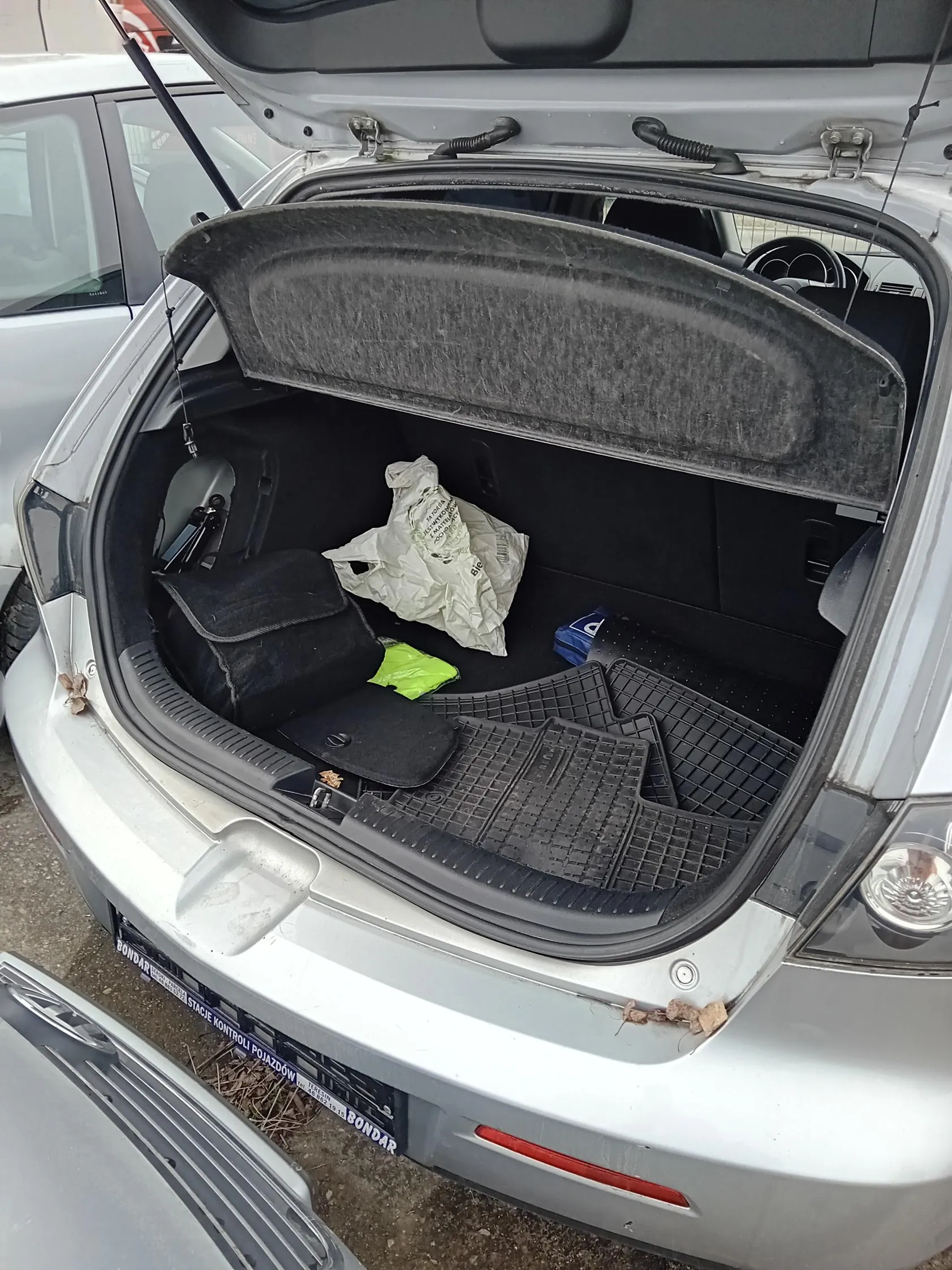 Otwarty bagażnik srebrnego samochodu z czarną torbą, białą torbą plastikową z niewyraźnymi napisami, zielonym przedmiotem oraz czarnymi gumowymi matami. Widoczna tablica rejestracyjna z napisem 'BONDAR' i 'STACJA KONTROLI POJAZDÓW'. W tle fragment innego pojazdu i nawierzchnia parkingowa.