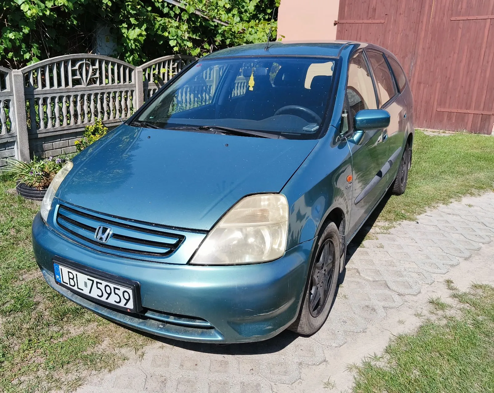 Niebieski samochód osobowy marki Honda zaparkowany na podjeździe z kostki brukowej i trawy. Pojazd ma polską tablicę rejestracyjną o numerze LBL 75959. W tle widoczny jest szary drewniany płot oraz brązowa brama.