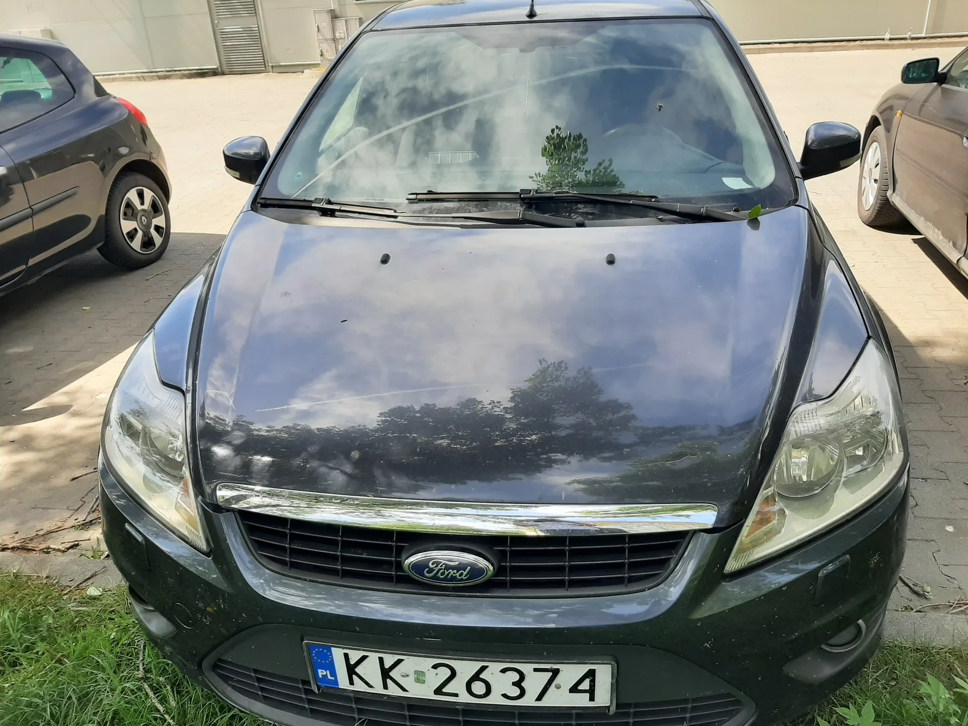 Czarny samochód marki Ford z przodu, z widocznym logo na atrapie chłodnicy i tablicą rejestracyjną PL KK 26374. Po obu stronach pojazdu znajdują się częściowo widoczne inne samochody: ciemnoniebieski po lewej i brązowy po prawej. Tło stanowi brukowana powierzchnia, trawa na dole kadru oraz budynek z wentylacją w tle. Na przedniej szybie odbijają się drzewa i chmury.