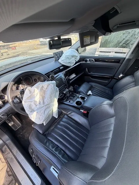 Wnętrze samochodu z rozwiniętymi poduszkami powietrznymi na kierownicy i po stronie pasażera. Czarne skórzane fotele, deska rozdzielcza z licznikami, kierownica po lewej stronie, centralna konsola z przyciskami i uchwytem na kubki. W tle widoczna droga z barierami i teren budowlany. W lusterku wstecznym widoczny żółty naklejony znak.