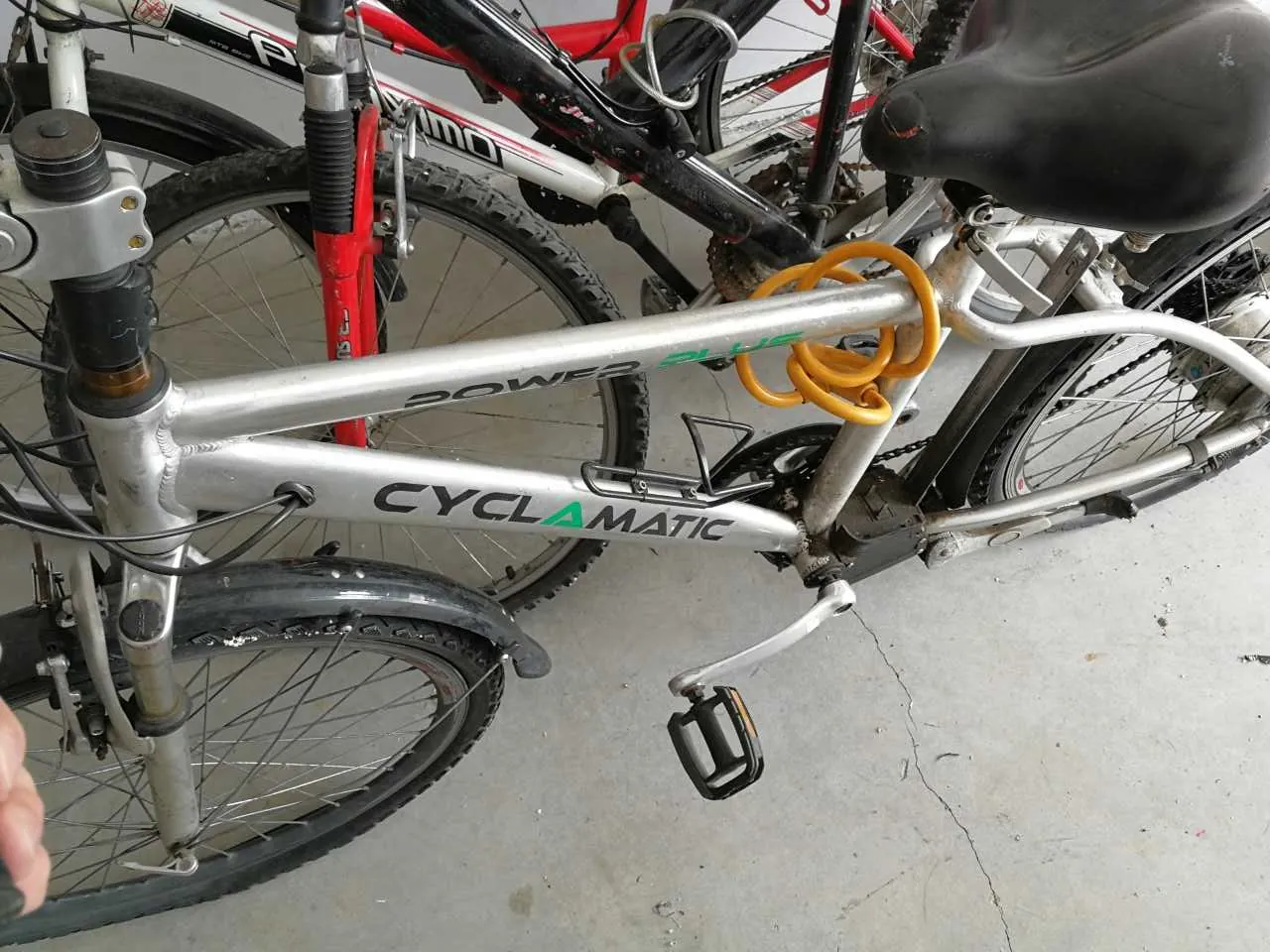 Zbliżenie na srebrny rower z napisem CYCLAMATIC na ramie, z żółtym zwiniętym kablem zabezpieczającym. W tle widoczne są części innych rowerów: czerwony i czarny. Podłoga z betonu z widocznymi pęknięciami. Na pierwszym planie widoczny pedał i koło przednie z oponą.