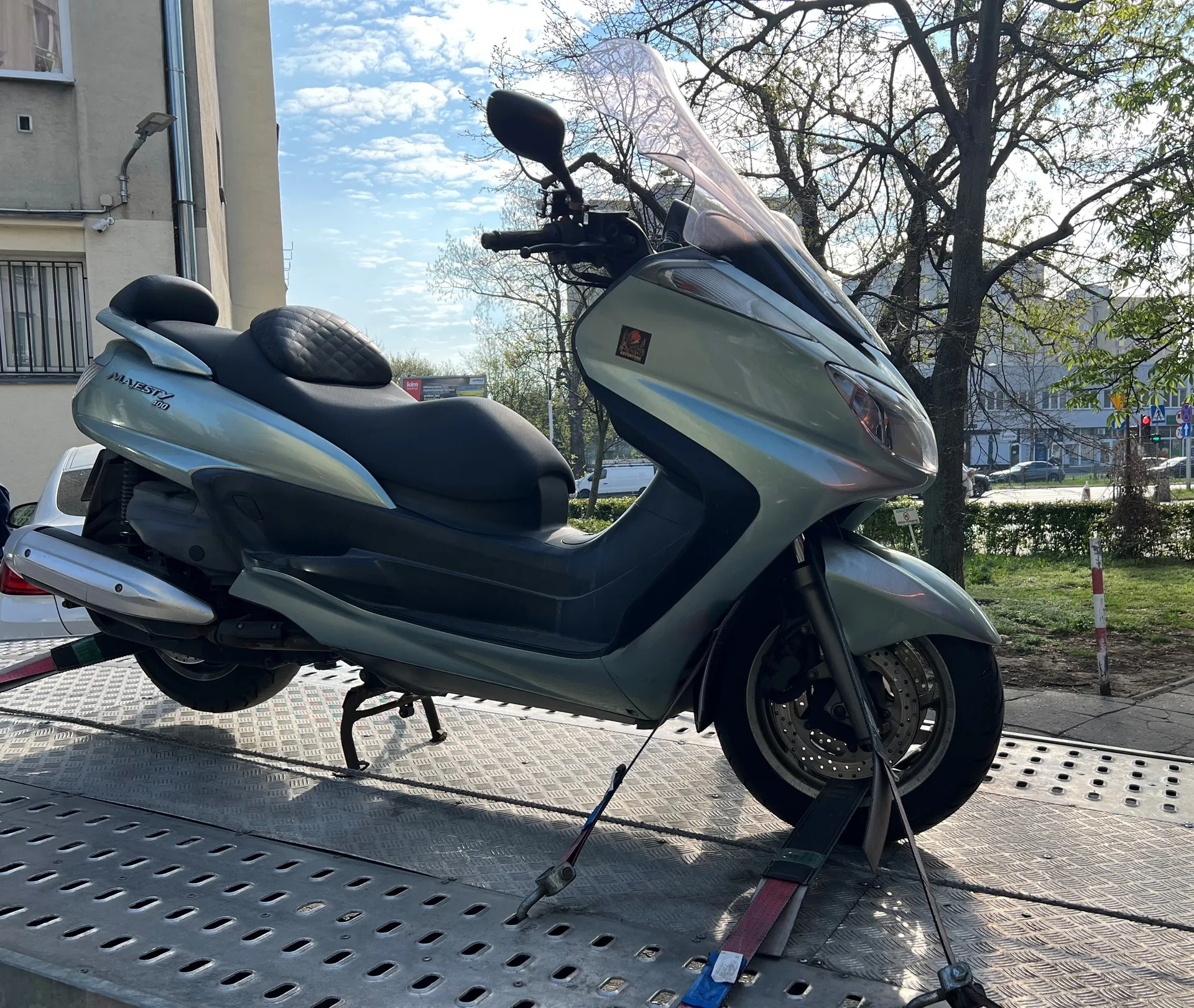 Srebrny skuter Honda Majesty 100 z czarnym siedzeniem i przednią osłoną, umocowany paskami na metalowej platformie z perforowanym wzorem. W tle budynek z oknami, drzewa, droga z pojazdami oraz niebo z rozproszonymi chmurami. Na boku skutera widoczny napis "MAJESTY 100".