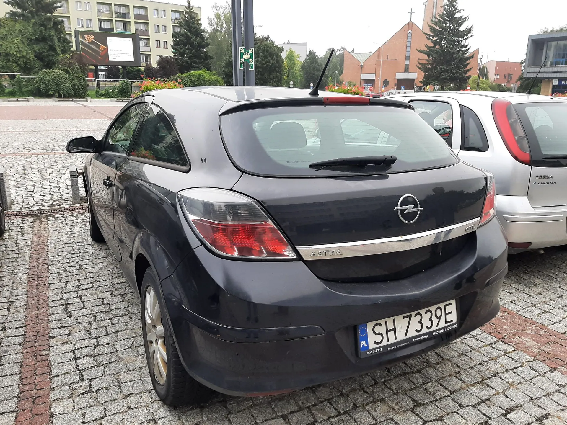 Czarny samochód marki Opel Astra zaparkowany na kostce brukowej. Widoczny jest tył i lewy bok pojazdu. Samochód ma polską tablicę rejestracyjną o numerze SH 7339E. W tle widać budynki mieszkalne, kościół oraz drzewa.