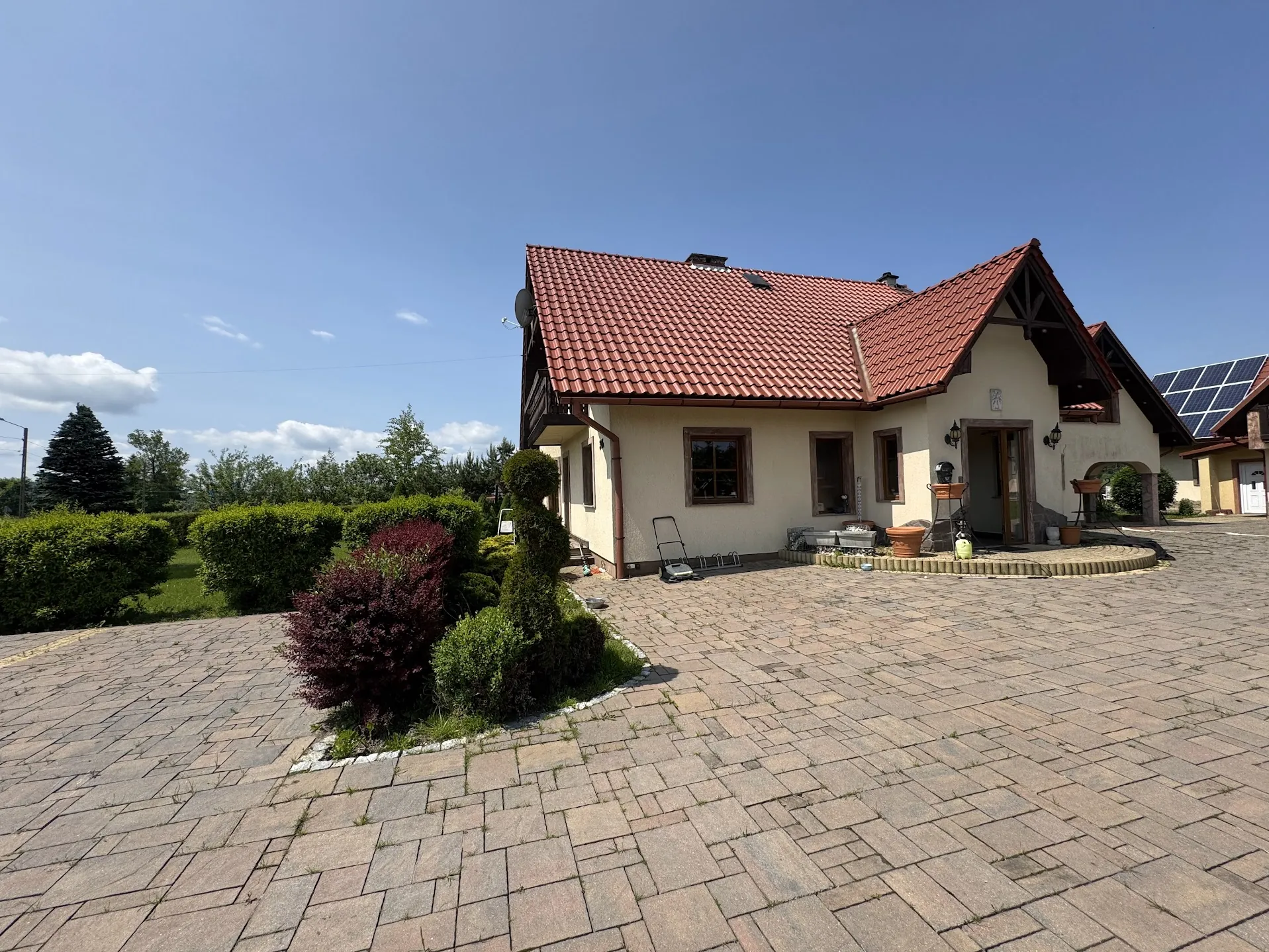 Dom jednorodzinny z czerwoną dachówką, jasnobrązowymi ścianami i brązowymi ramami okiennymi. Przed domem brukowany dziedziniec z kamiennymi płytami. Wokół domu zielone krzewy, w tym ciemnoczerwony krzew i wysoki, uformowany krzew. Po prawej stronie widoczna część innego budynku z panelami słonecznymi na dachu. Na tle błękitne niebo z kilkoma chmurami oraz drzewa. Na dziedzińcu stoją donice, wózek ogrodniczy i inne przedmioty. Horyzont jest poziomy, a budynki są prawidłowo ustawione.