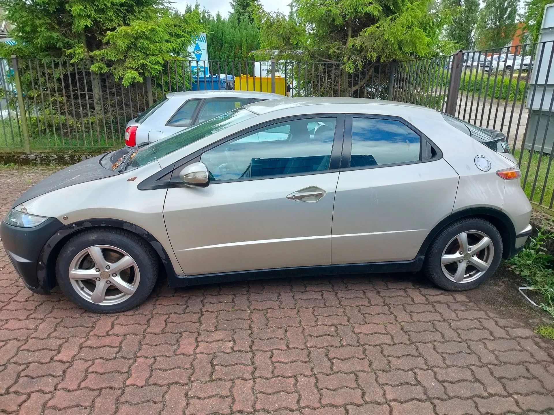 Srebrny samochód osobowy marki Honda Civic w nadwoziu hatchback, zaparkowany bokiem na kostce brukowej. Auto ma ciemną maskę i czarne progi. W tle widoczny jest drugi, jasny samochód, metalowy płot oraz zielone drzewa i krzewy.
