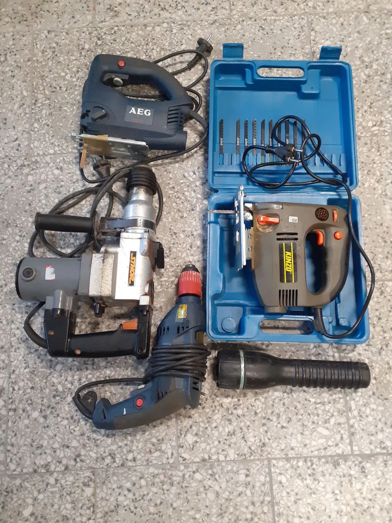Zestaw narzędzi elektrycznych ułożonych na jasnej, ziarnistej posadzce. W górnej części znajduje się wyrzynarka marki AEG. Po lewej stronie widoczna jest duża wiertarka udarowa z dodatkowym uchwytem. Na dole znajduje się wiertarko-wkrętarka oraz latarka. Po prawej stronie znajduje się otwarta niebieska walizka narzędziowa, w której umieszczona jest wyrzynarka marki Kinzo wraz z zestawem brzeszczotów.