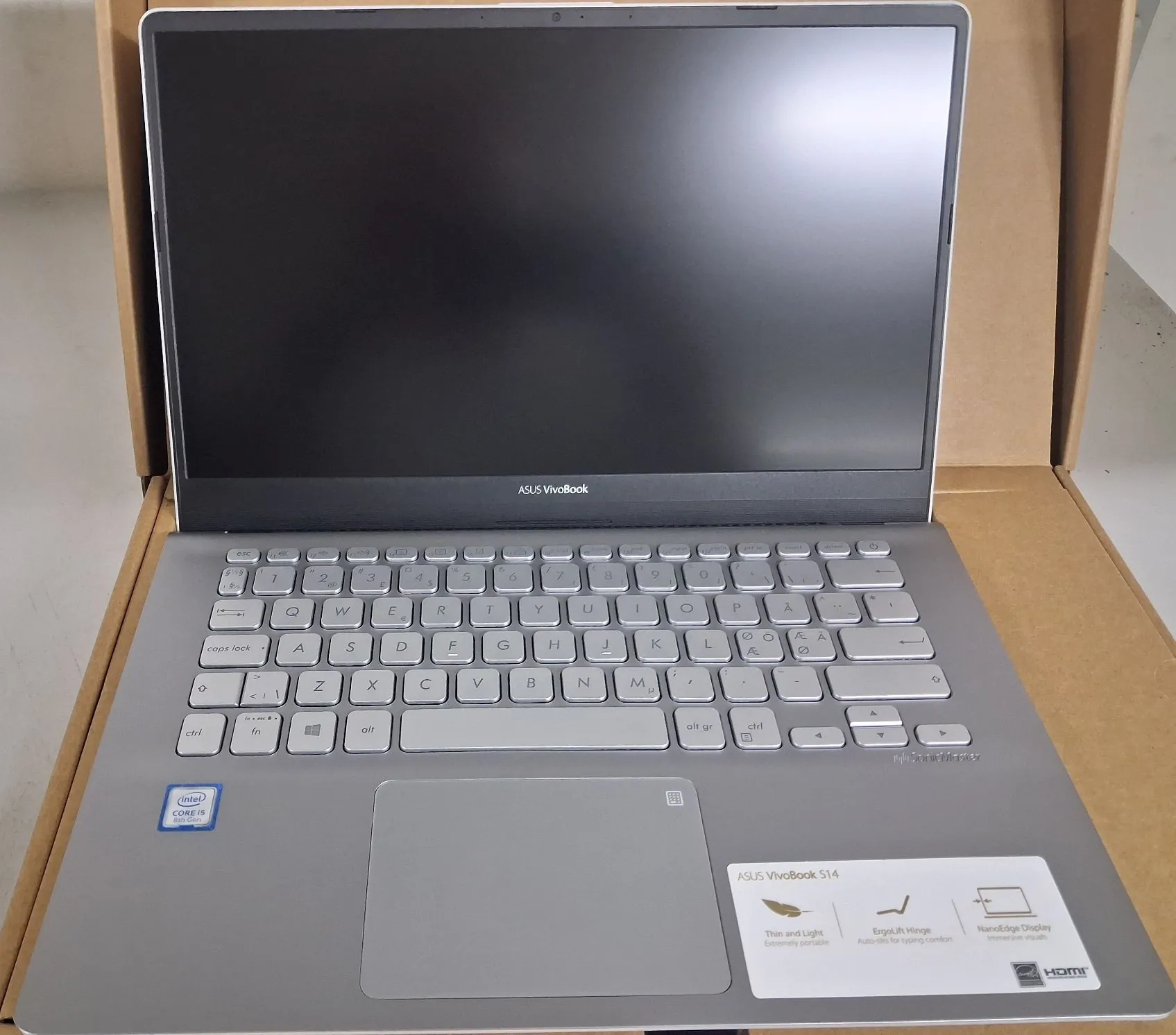 Srebrny laptop ASUS VivoBook S14 otwarty i umieszczony w brązowym kartonowym opakowaniu. Widoczna jest klawiatura z podświetleniem, gładzik oraz ekran w kolorze czarnym. Na obudowie poniżej klawiatury znajdują się naklejki: logo Intel Core i5, napis ASUS VivoBook S14 oraz ikony prezentujące cechy urządzenia: Thin and Light, ErgoLift Hinge, NanoEdge Display i złącze HDMI.