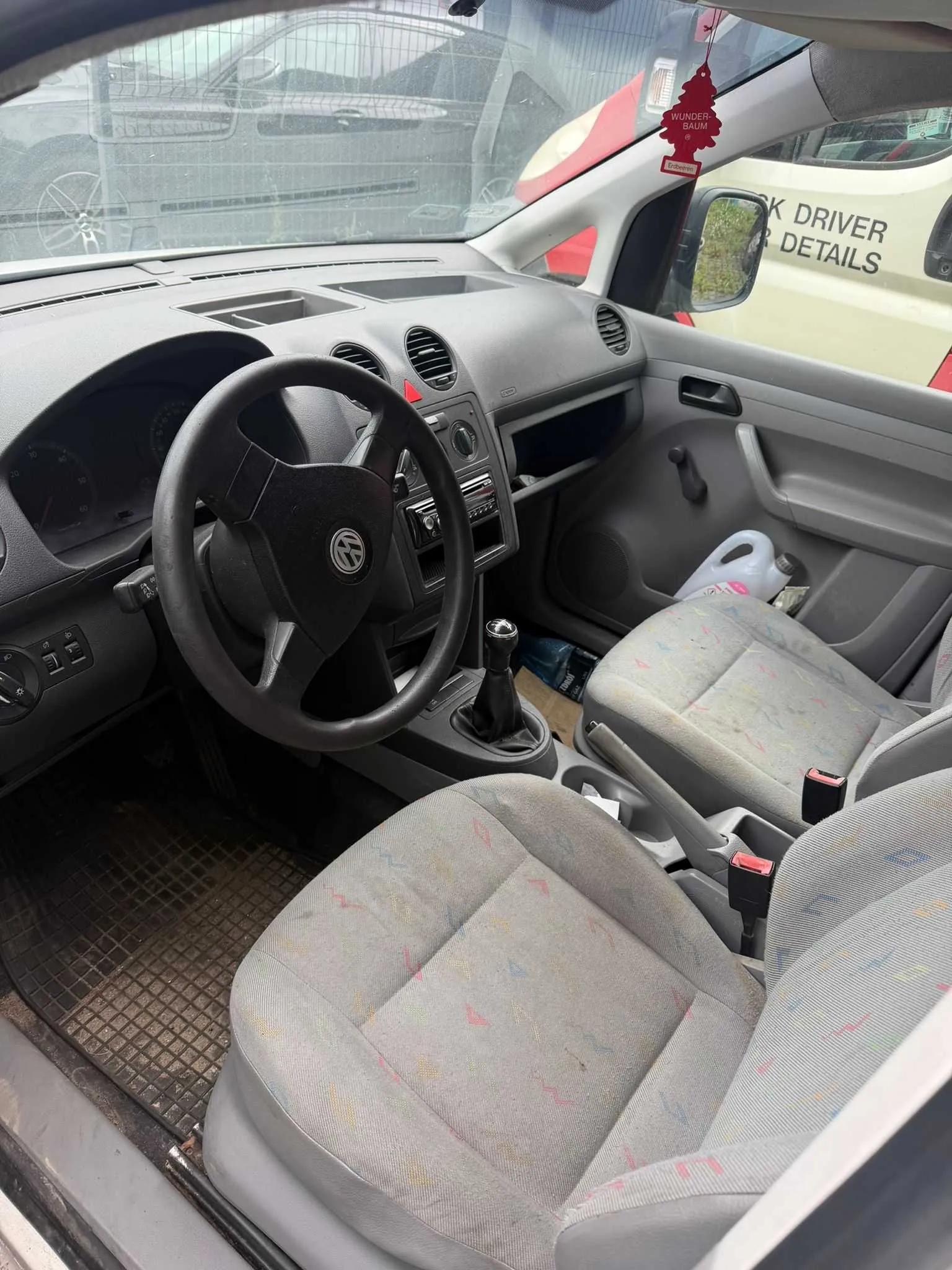 Wnętrze samochodu widziane z perspektywy pasażera. Widoczna jest kierownica z logo Volkswagena, deska rozdzielcza, lewy fotel kierowcy oraz prawy fotel pasażera w szarej, wzorzystej tapicerce. Na podłodze znajduje się gumowa mata. Przez przednią szybę widoczny jest fragment innego, czerwonego samochodu z naklejką w kształcie czerwonego ośmiokąta z napisem 'WUNDER BAUM' oraz fragment białego pojazdu z napisem 'DRIVER DETAILS'.