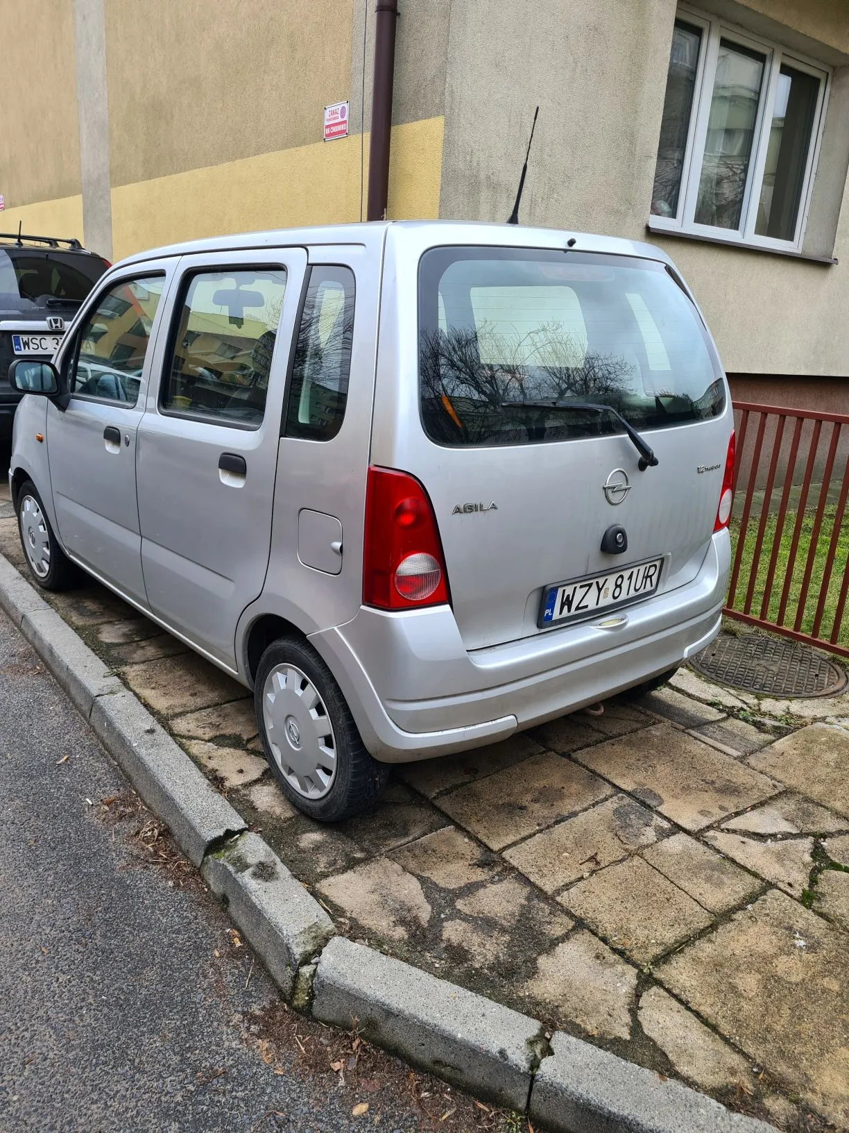 Srebrny samochód Opel Agila zaparkowany przy budynku. Widoczna tablica rejestracyjna WZY 81UR. Budynek z oknem, czerwoną balustradą i żółto-beżową elewacją. Na chodniku z kamieni widoczne są ślad po kałużach. Za samochodem częściowo widoczny inny pojazd z tablicą rejestracyjną. Na ścianie budynku mały czerwono-biały znak.