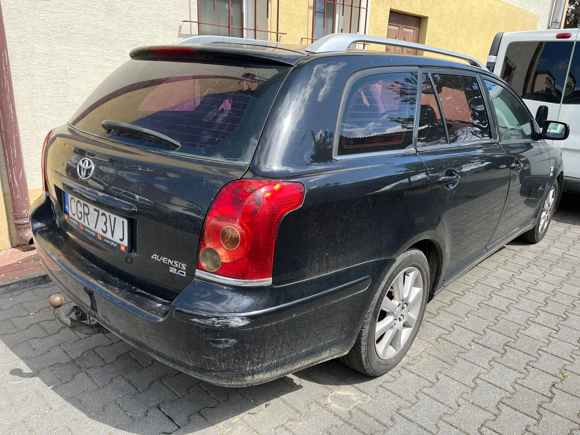 Czarny samochód marki Toyota Avensis 2.0 w nadwoziu kombi, zaparkowany na kostce brukowej. Widoczny jest tył i prawy bok pojazdu. Samochód ma polską tablicę rejestracyjną o numerze CGR 73VJ. Na tylnym zderzaku widoczne są zarysowania, a pod nim zamontowany jest hak holowniczy.