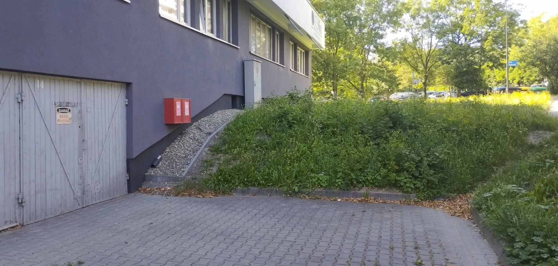 Zdjęcie przedstawia fragment szarego budynku z zamkniętymi, jasnymi drzwiami garażowymi po lewej stronie. Na drzwiach znajduje się mała tabliczka z napisem „GARAŻ NIE wjeżdżać”. Obok drzwi, na ścianie budynku, zamontowana jest czerwona skrzynka przeciwpożarowa. Przed budynkiem znajduje się obszar z kostki brukowej, a obok niego nasyp z kamieni oraz gęsty pas zieleni z trawą i krzewami. W tle widoczne są drzewa i fragmenty zaparkowanych samochodów.