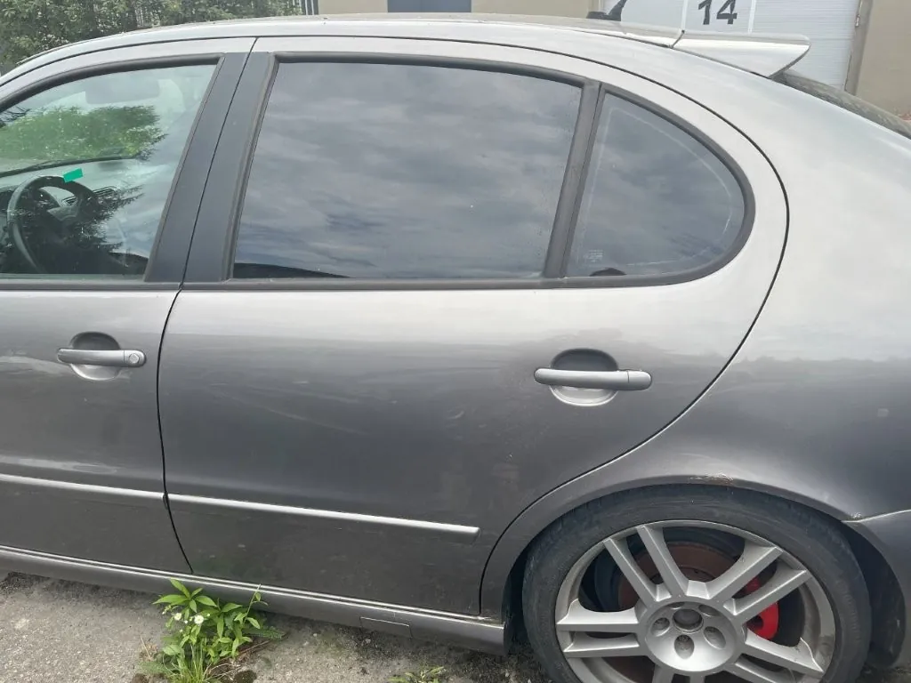 Boczny widok szarego samochodu osobowego typu hatchback. Widoczne są przednie i tylne drzwi z klamkami, przyciemniane tylne szyby oraz tylne koło z wieloramienną srebrną felgą i czerwonym zaciskiem hamulcowym. W tle w górnej części widoczny jest fragment budynku z numerem 14.