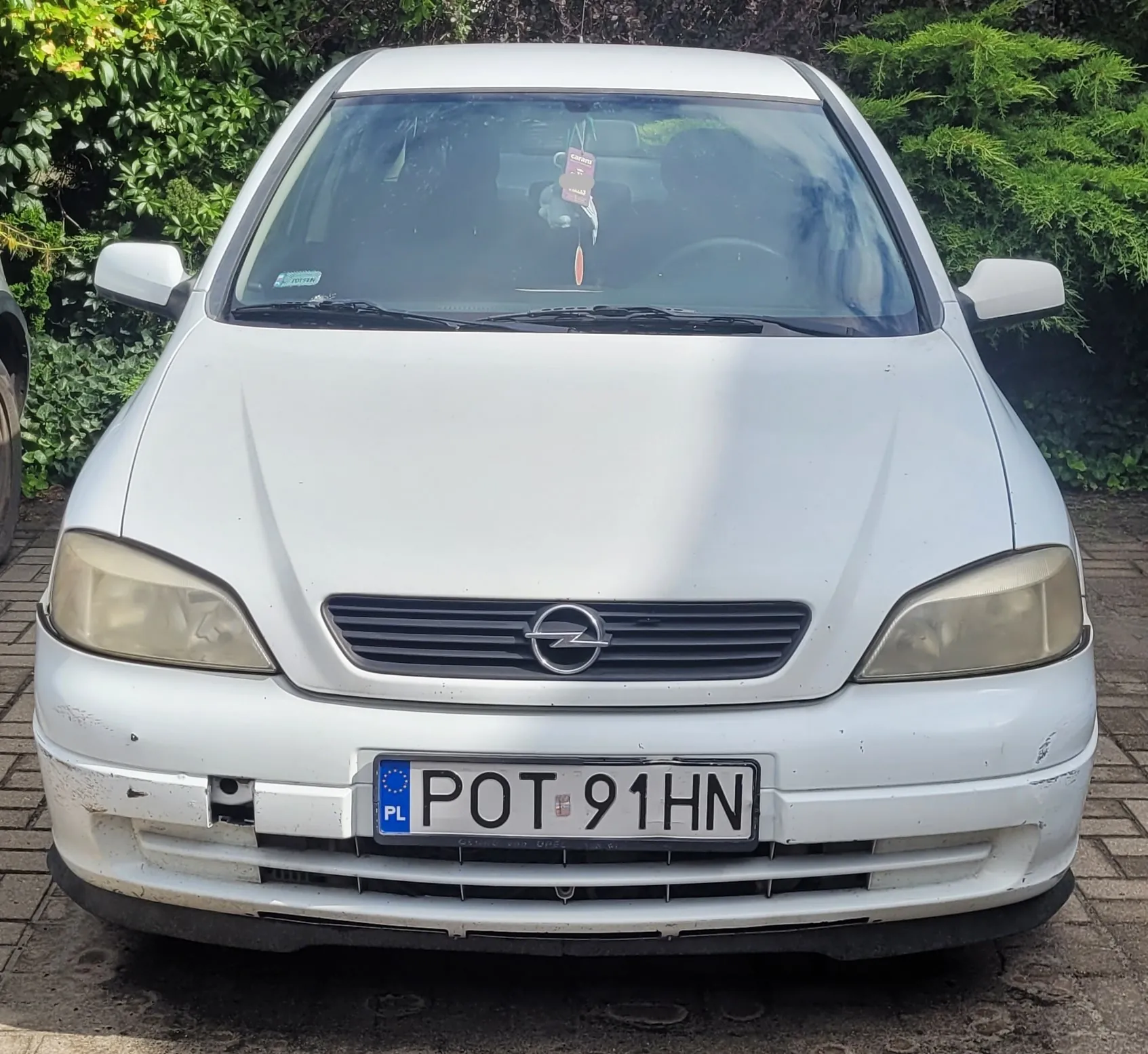 Widok z przodu białego samochodu marki Opel. Pojazd ma żółtawe reflektory, czarny grill z logo producenta oraz polską tablicę rejestracyjną o numerze POT 91HN. Samochód stoi na kostce brukowej na tle zielonych krzewów.