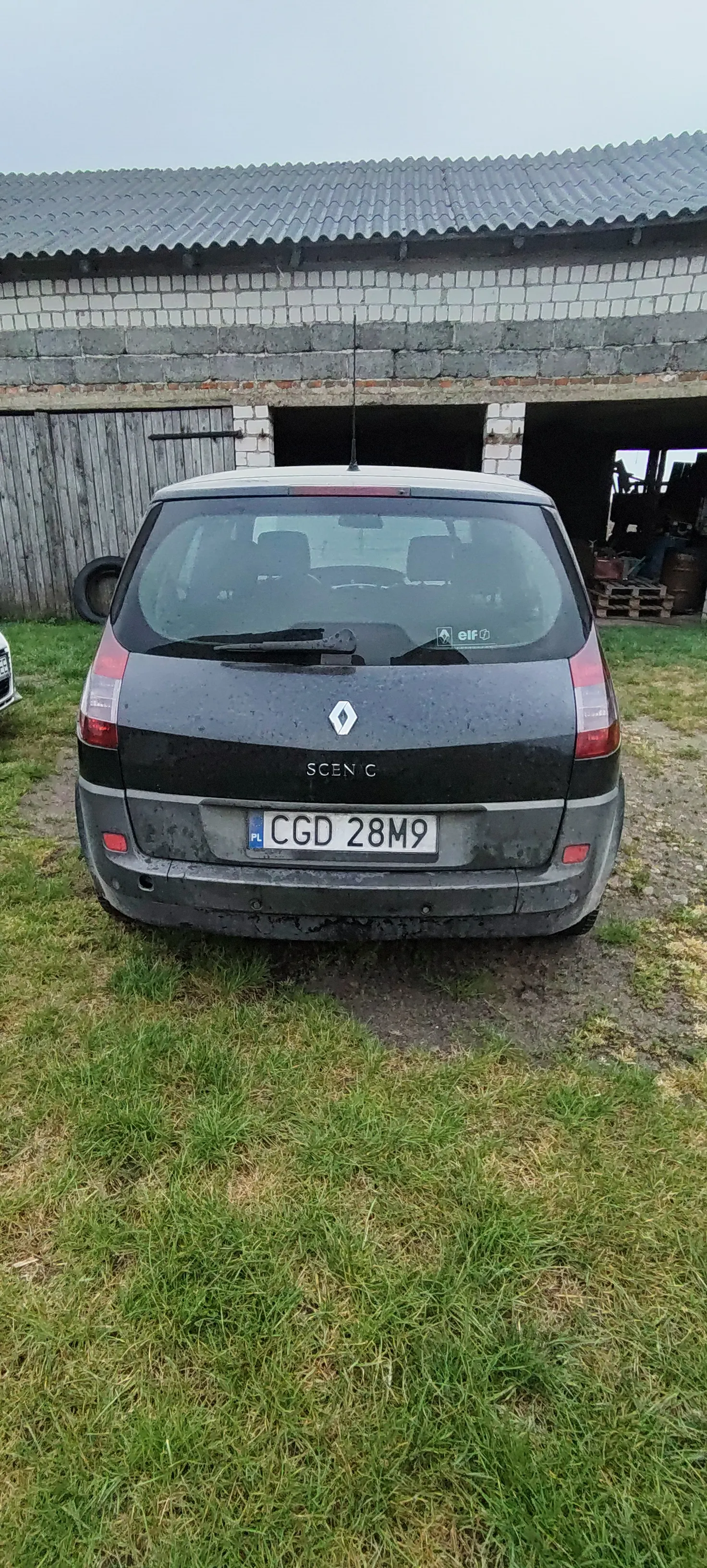 Czarny samochód Renault Scenic z tablicą rejestracyjną CGD 28M9 zaparkowany na podwórku. Po lewej stronie widoczny budynek z ceglaną ścianą i dachem z blachy falistej, w tle otwarte pomieszczenie z narzędziami. Podłoga składa się z trawy i żwiru, niebo jest pochmurne. Na tylnym szybie samochodu widoczne naklejki, w tym logo marki i napis SCENIC.