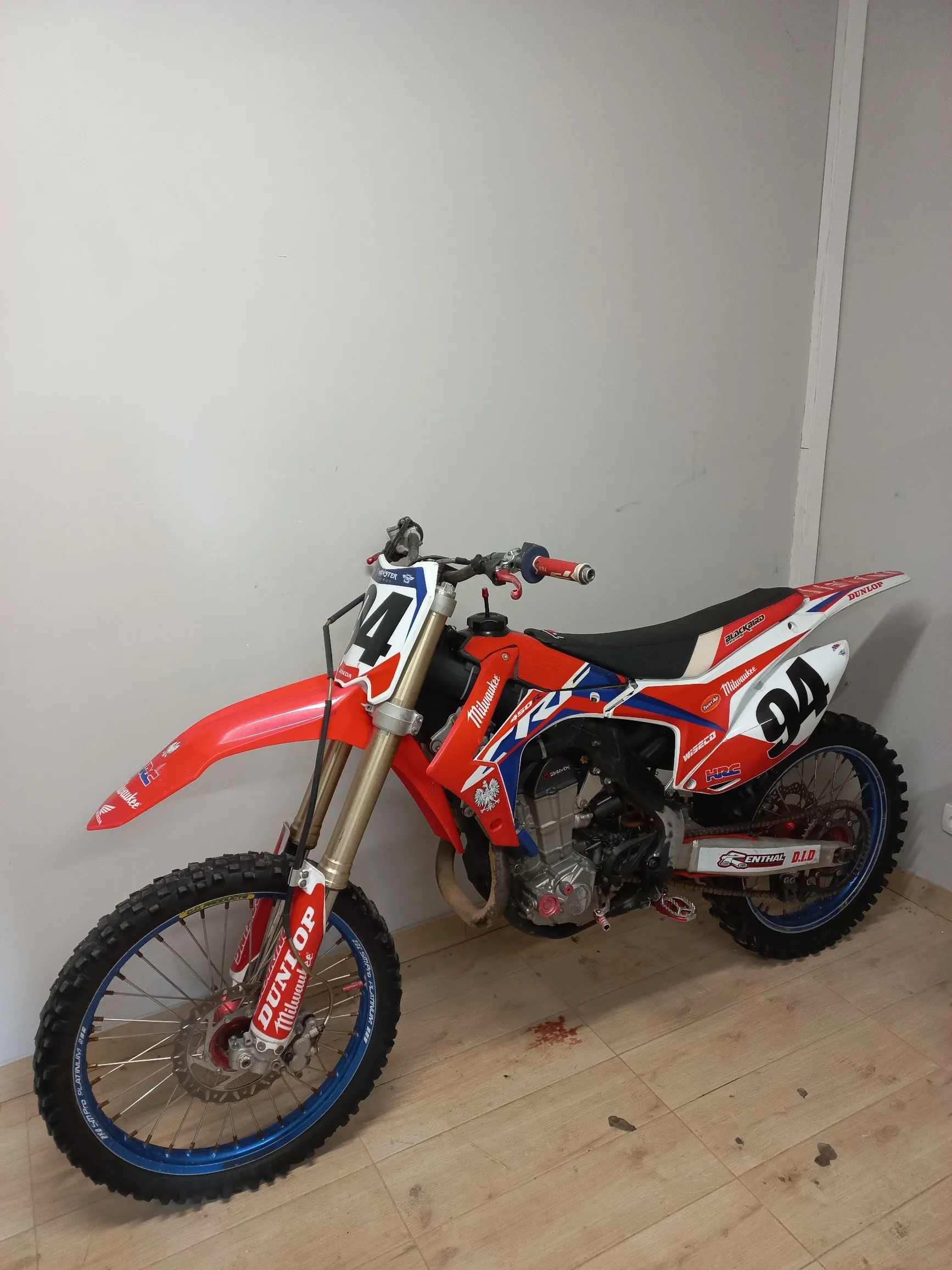 Motocykl crossowy w kolorach czerwonym, białym i niebieskim, ustawiony bokiem na jasnej drewnianej podłodze na tle białej ściany. Motocykl posiada numer 94 na tylnym błotniku i numer 4 na przedniej owiewce. Na owiewkach widoczne są logotypy marek: Honda, HRC, Dunlop oraz Renthal. Koła mają niebieskie obręcze i terenowe opony.