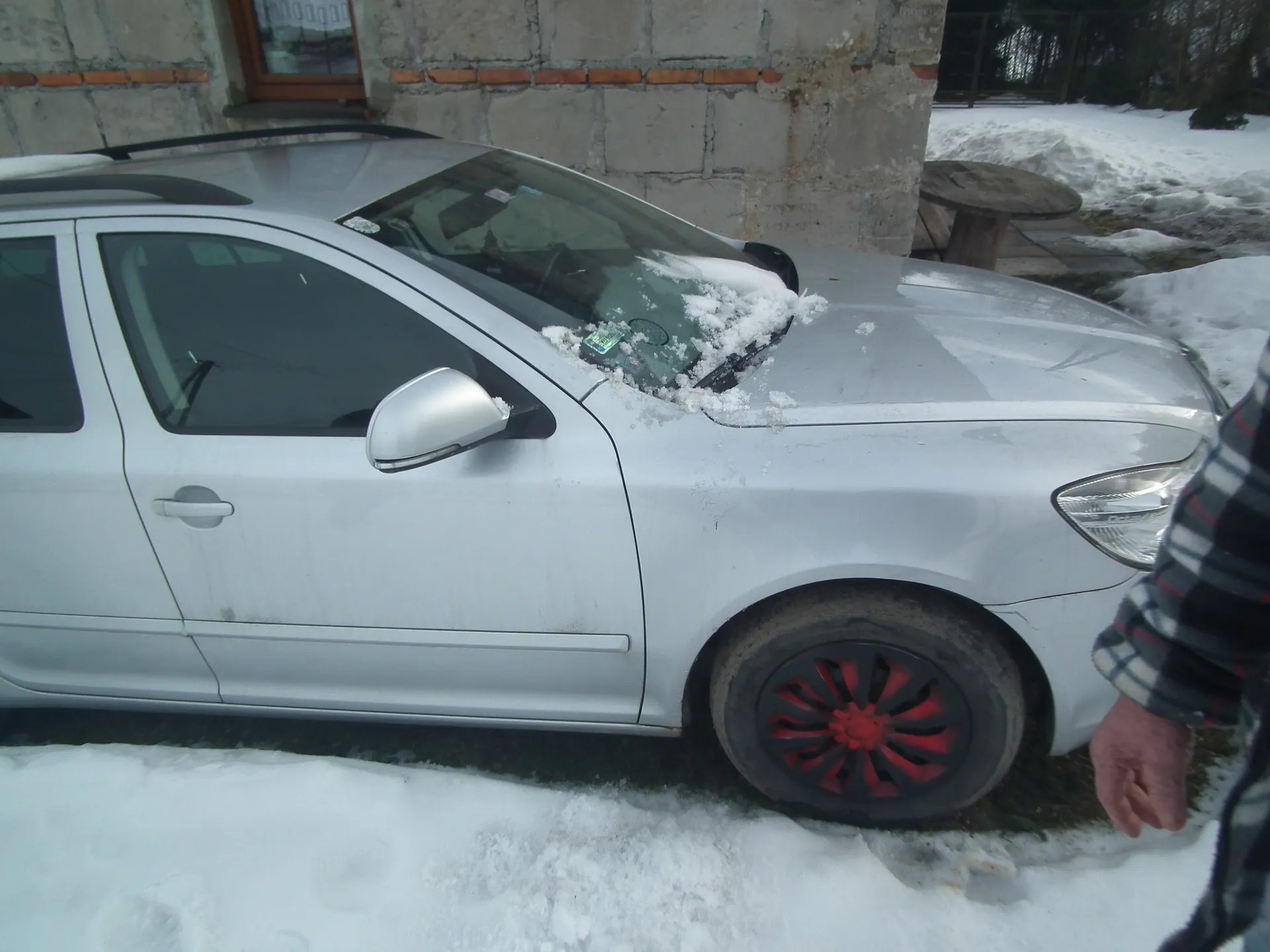 Srebrny samochód osobowy zaparkowany na śniegu przed budynkiem z betonowych bloczków i czerwonych cegieł. Na masce i przedniej szybie widoczny śnieg. Koło przednie z czerwono-czarną felgą. Po prawej stronie fragment ręki w kratowanym swetrze. W tle stół ogrodowy i śnieżna powierzchnia.
