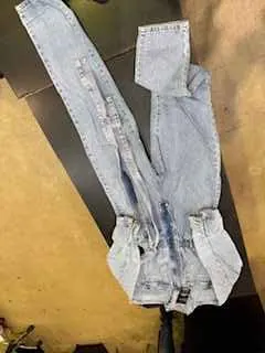 Jasnoniebieskie dżinsowe kombinezony leżące płasko na powierzchni. Kombinezony mają zamknięty na suwak, kieszenie i rozłożone nogawki. Tło składa się z ciemnego stołu i jasnej podłogi.