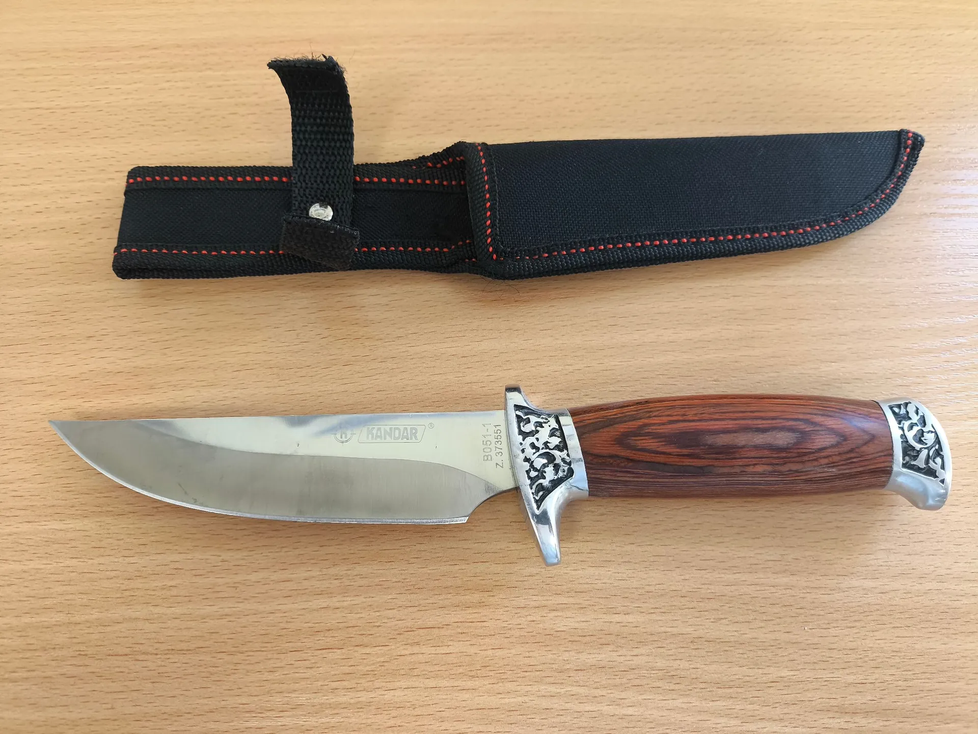 Nóż z metalowym ostrzem i drewnianą rękojeść leżący na jasnym, drewnianym blacie. Na ostrzu widnieje napis 'KANDAR' oraz oznaczenia 'B0511-1 Z. 373551'. Obok noża znajduje się czarna, materiałowa pochwa z czerwonymi przeszyciami.
