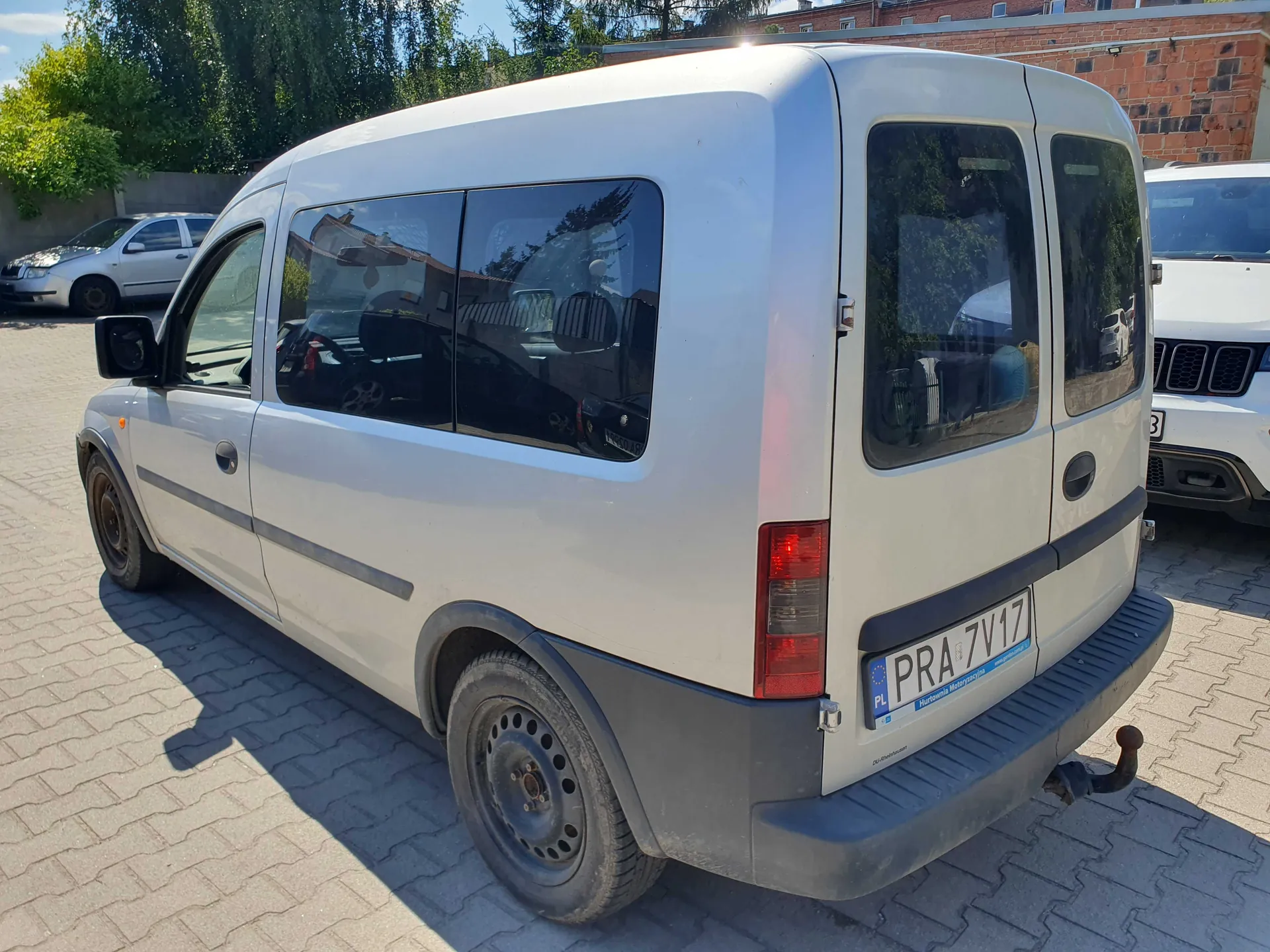 Biały samochód dostawczy typu van zaparkowany na kostce brukowej. Widoczny jest tył i lewy bok pojazdu. Samochód ma czarne plastikowe zderzaki i listwy boczne oraz stalowe felgi. Z tyłu znajduje się tablica rejestracyjna o numerze PRA 7V17. W tle widoczny jest fragment innego białego samochodu oraz ceglany budynek i drzewa.