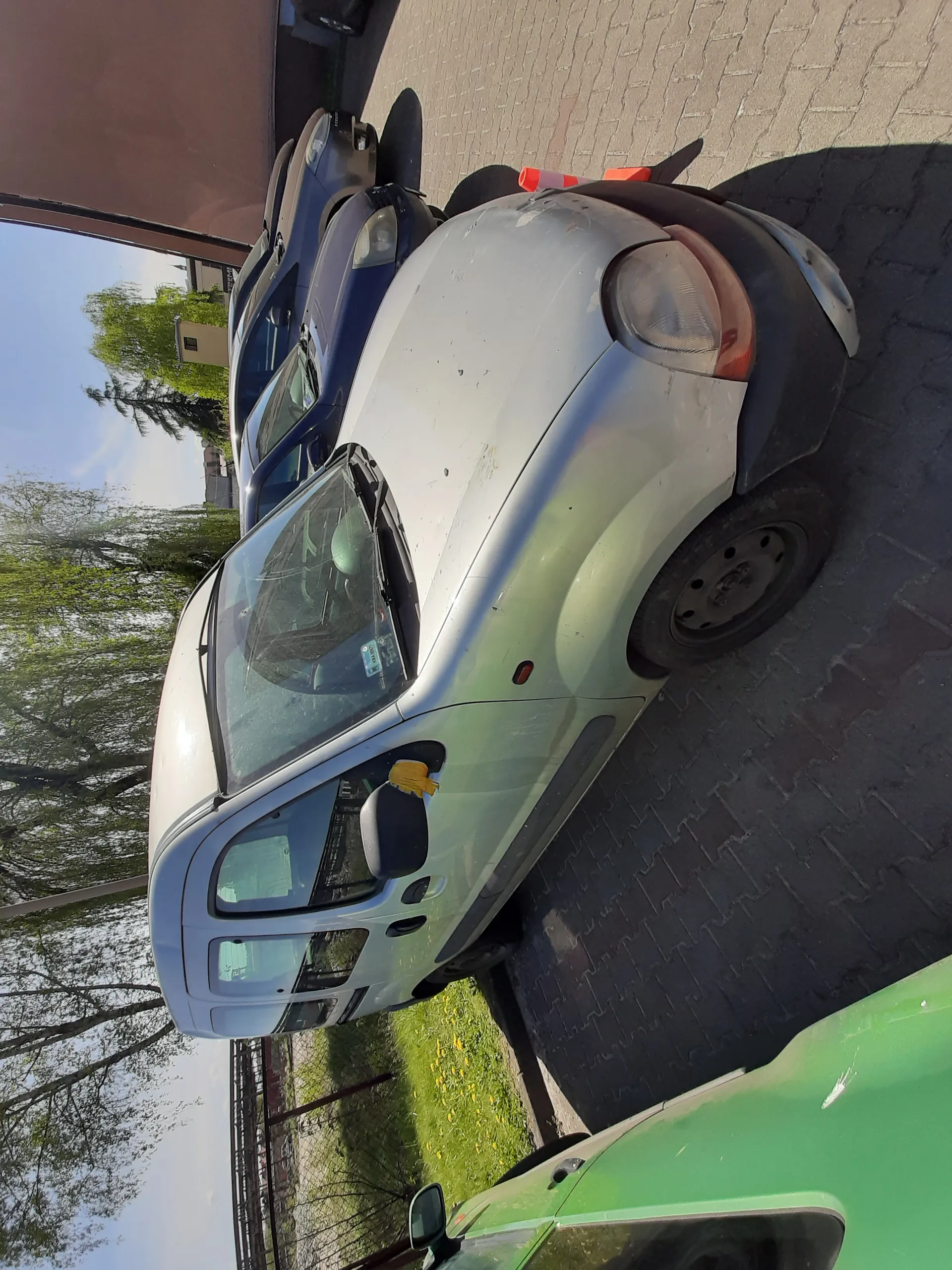 Srebrny furgonetka zaparkowana na brukowanym placu parkingowym. W tle widoczne są inne samochody, zielony samochód po lewej stronie, trawa, drzewa i budynek. Na lusterku bocznym furgonetki znajduje się żółta szmatka. Na placu parkingowym stoją pomarańczowe stożki.