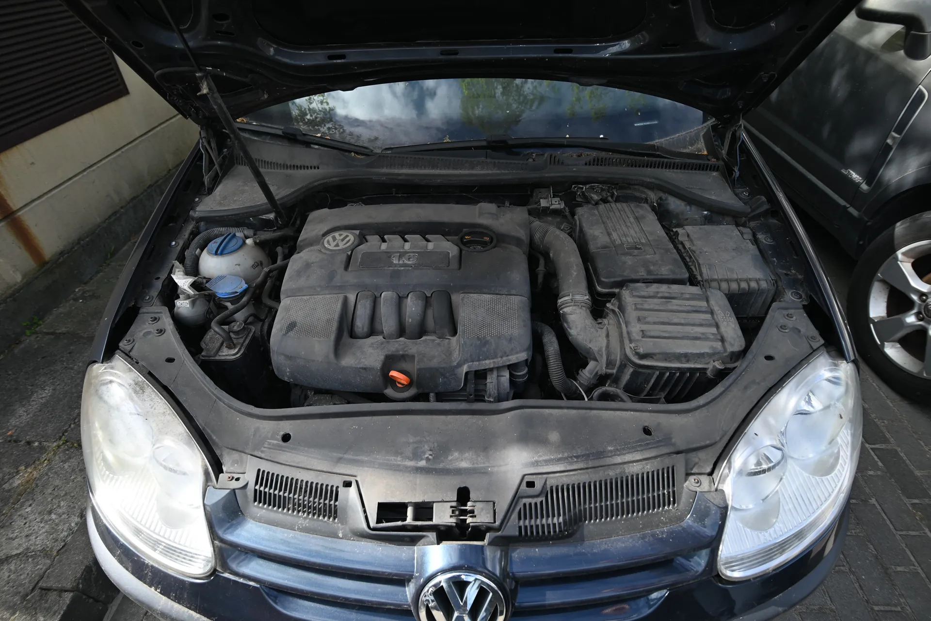 Samochód marki Volkswagen z otwartą maską, ukazujący silnik z widocznym logo VW i oznaczeniem 1.6. W silniku widoczne są elementy takie jak zbiornik z niebieską pokrywką, przewody i czarne osłony. Samochód stoi na brukowanej powierzchni obok budynku z betonową ścianą i ciemnym oknem. Po prawej stronie częściowo widoczny jest inny pojazd. Przednia część samochoda z reflektorami i chłodnikiem jest wyraźnie widoczna.