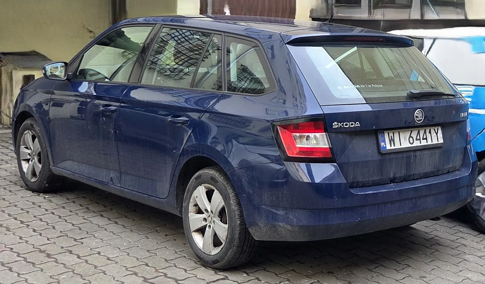Niebieski samochód marki ŠKODA typu kombi zaparkowany na brukowanej powierzchni. Widoczny tył pojazdu z tablicą rejestracyjną PL WI 6441Y oraz logo marki. Na tylnej szybie napis „ŠKODA No.1 w Polsce”. Samochód ma brudne boki i koła, w tle fragment budynku oraz częściowo widoczny inny pojazd. Szyby boczne osłonięte siatką przeciwsłoneczną.