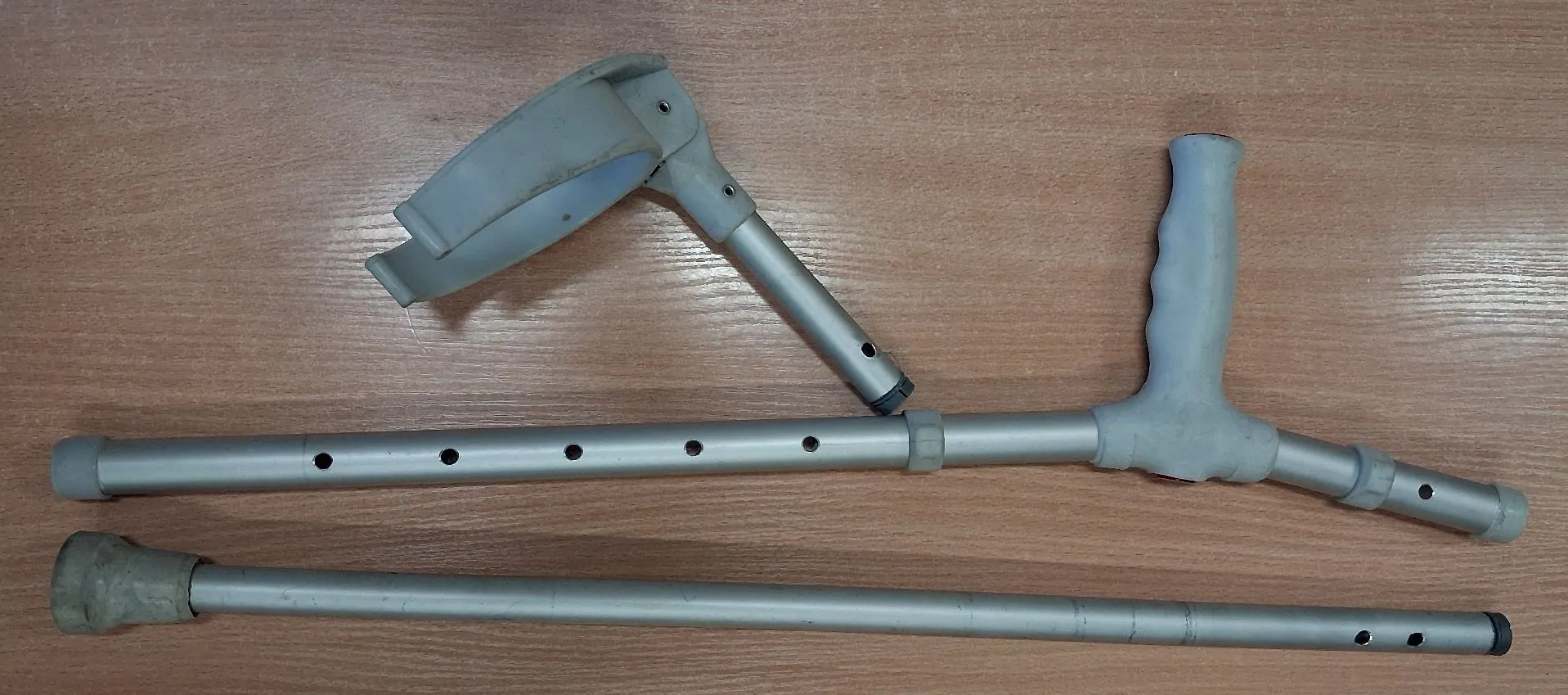 Rozłożone części metalowych kul kulawych w kolorze srebrnym, ułożone na brązowym blacie o strukturze drewna. Widoczny jest uchwyt przedramionny, rączka oraz dwie rurki z otworami do regulacji wysokości.
