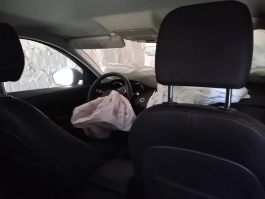 Wnętrze samochodu widziane z tylnego siedzenia. Widoczne czarne oparcia przednich foteli, kierownica, zdeformowana różowa poduszka powietrzna po stronie kierowcy oraz fragmenty zewnętrzne przez przednią szybę: kamienna ściana i biały materiał przykrywający przedmiot poza samochodem. Wnętrze jest ciemne, a zewnętrzne elementy są słabo oświetlone.