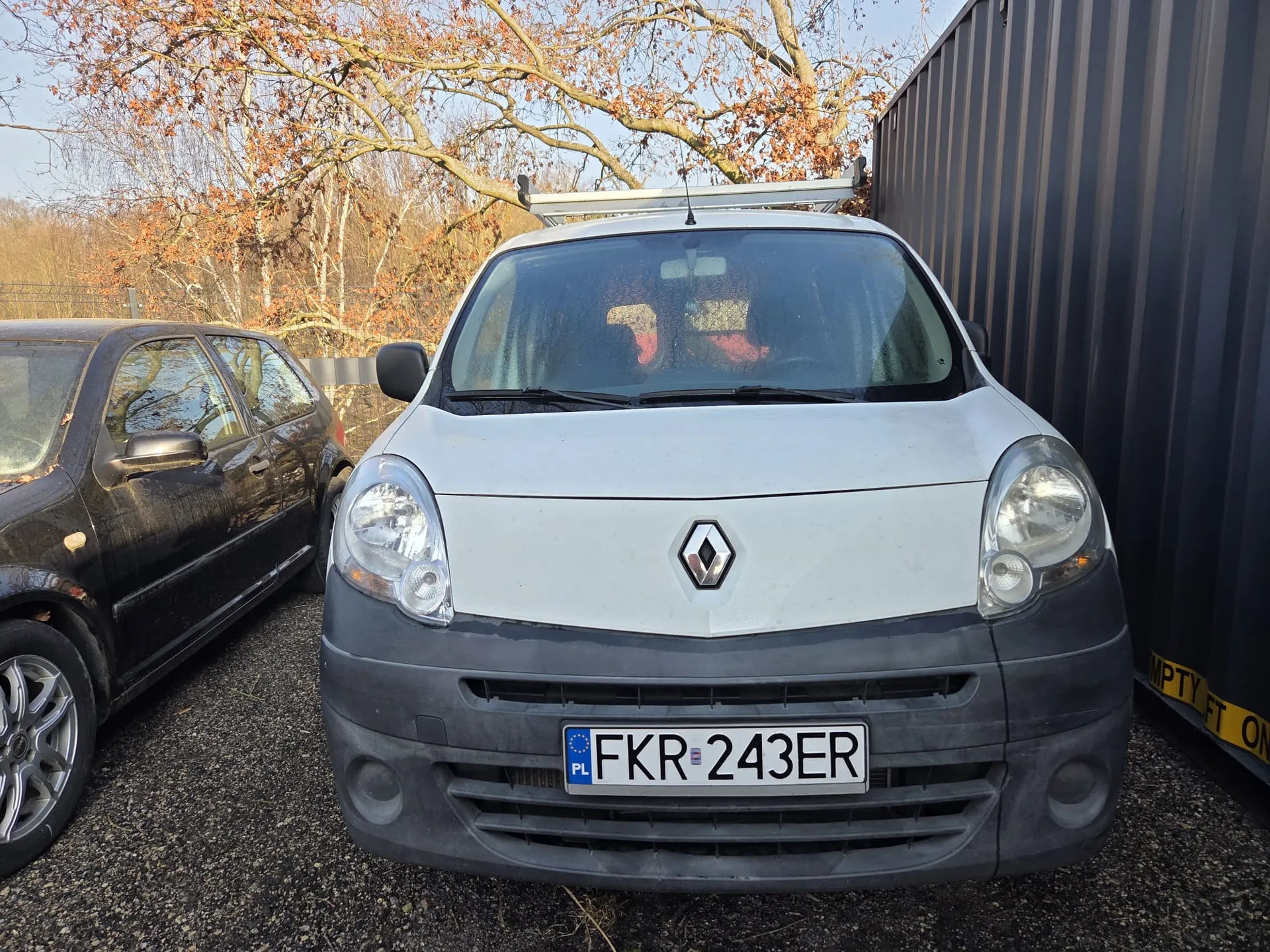 Biały samochód dostawczy marki Renault z tablicą rejestracyjną FKR 243ER (PL) na pierwszym planie. Po lewej stronie czarny samochód osobowy. W tle drzewa z brązowymi liśćmi i ciemny kontener z częściowo widocznym napisem 'EMPTY FT ON'. Samochód ma dachowy uchwyt, a przednia szyba odbija sylwetki osób.