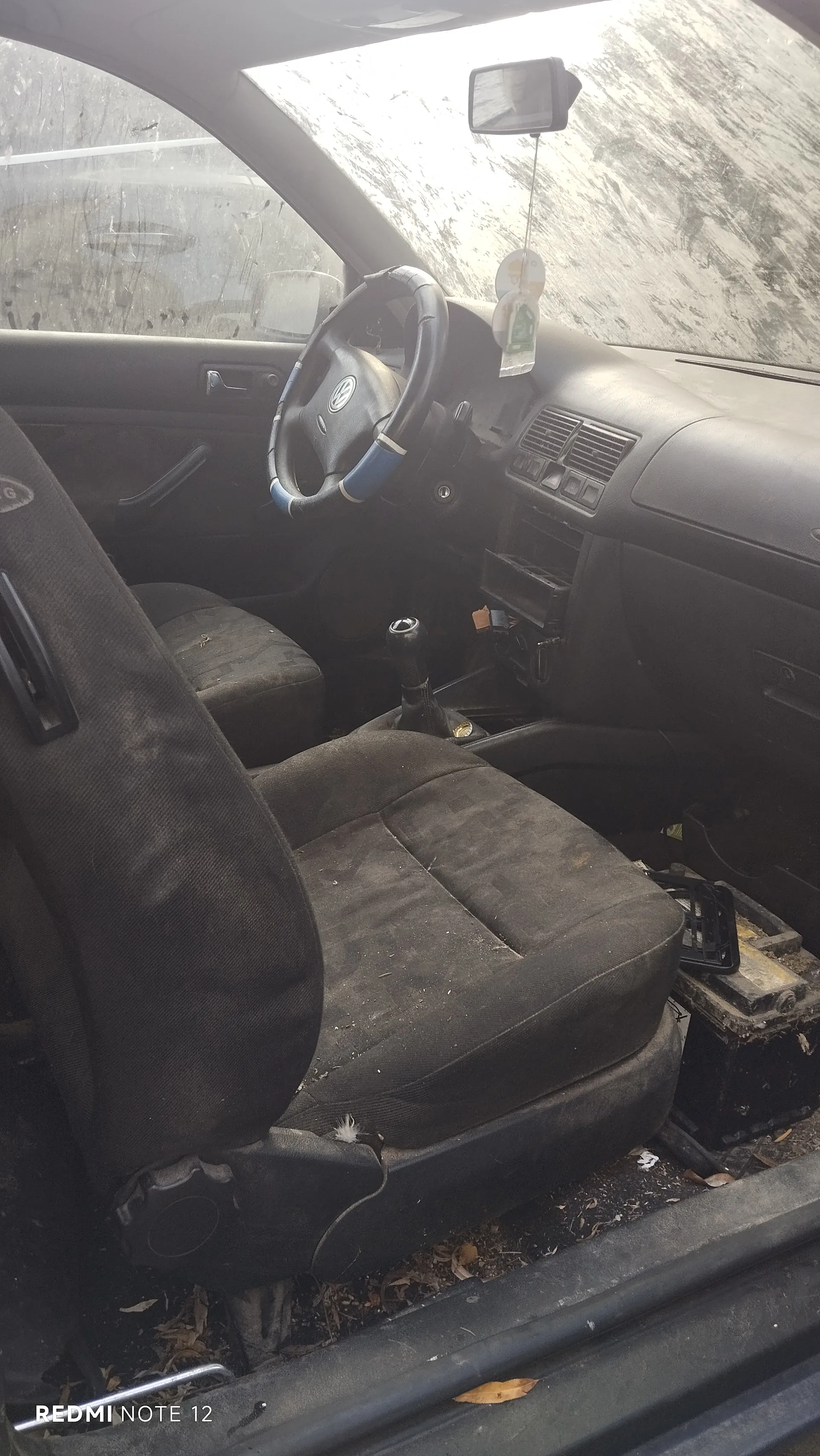 Wnętrze samochodu marki Volkswagen z widocznym kierownicą z logo VW, brudnymi siedzeniami, dźwignią zmiany biegów i zanieczyszczonym deski rozdzielczej. Na lusterku wstecznym wiszą ozdoby. Przez przednią szybę widać zanieczyszczoną powierzchnię zewnętrzna. W lewym dolnym rogu widoczny napis REDMI NOTE 12. Podłoga wewnątrz pojazdu pokryta jest śmieciem i liśćmi.