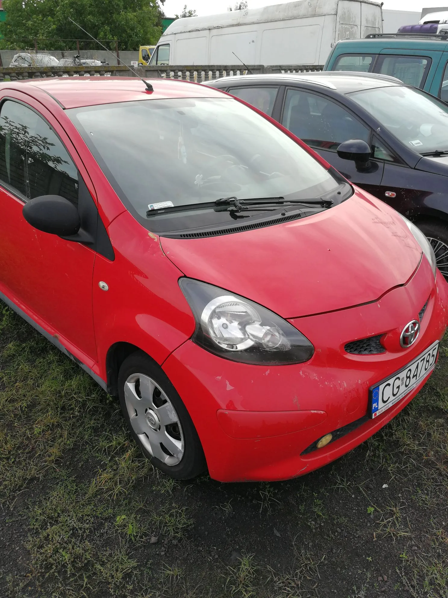 Czerwony samochód osobowy marki Toyota zaparkowany na nieutwardzonym terenie. Widoczny jest przód pojazdu z tablicą rejestracyjną o numerze CG 84785. W tle znajdują się inne samochody, w tym biały furgon oraz ciemny samochód osobowy.