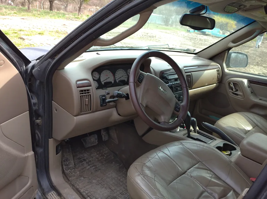 Wnętrze samochodu marki Jeep z otwartą drzwiami kierowcy. Beżowa tapicerka, kierownica z logo Jeep, deska rozdzielcza z licznikami, radio i klimatyzacją. Widoczne są brudne dywany podłogowe oraz zewnętrzny widok na drogę ziemną i drzewa za przednią szybą.
