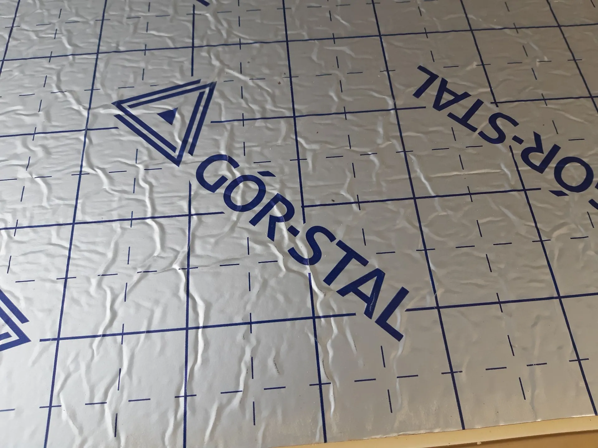 Zbliżenie na srebrną, pogniecioną folię ochronną z nadrukowaną niebieską siatką linii oraz dużym niebieskim napisem „GÓR-STAL” wraz z logo w kształcie trójkąta.