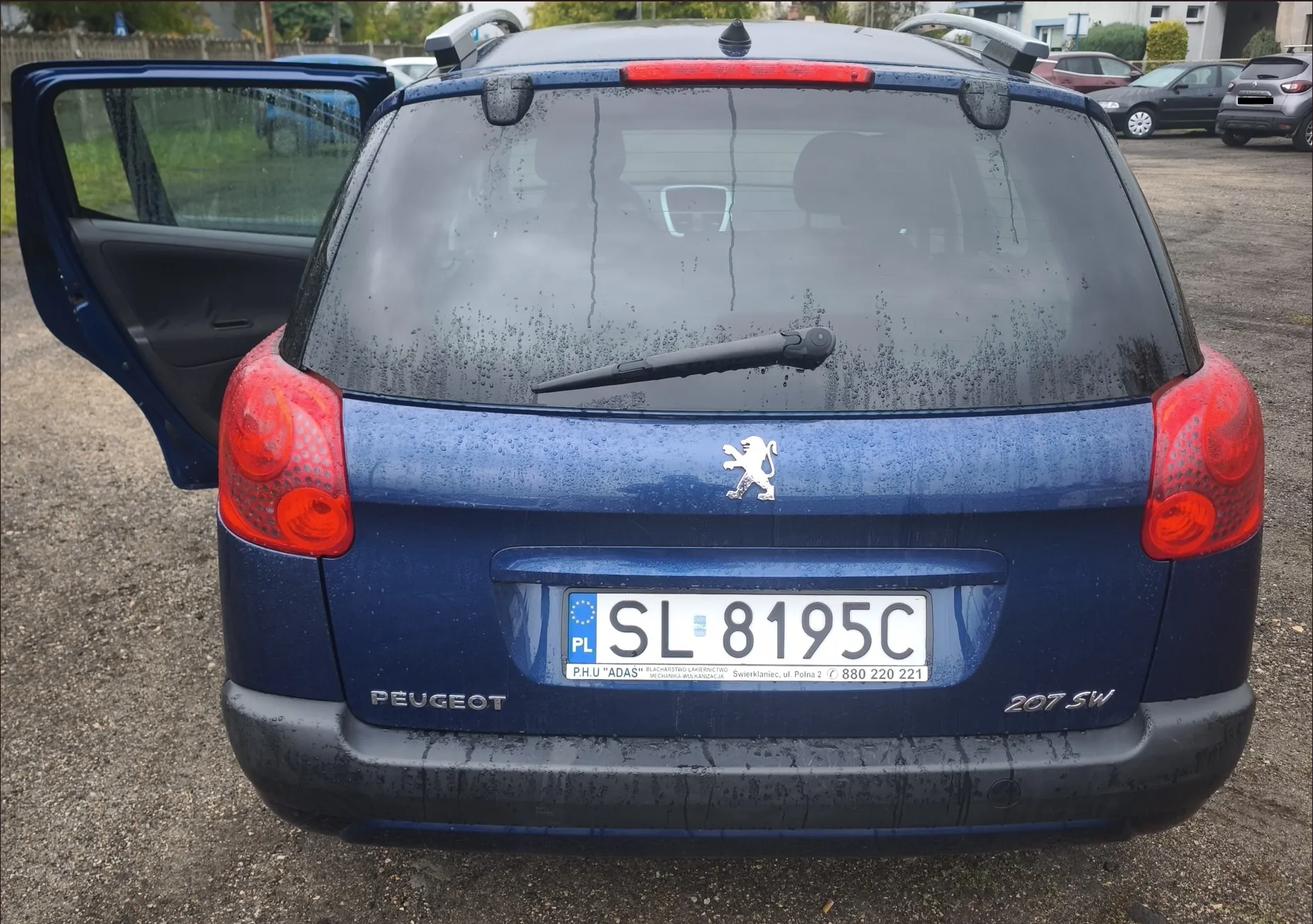 Tylny widok niebieskiego samochodu marki Peugeot 207 SW z otwartymi tylnymi drzwiami po lewej stronie. Samochód jest mokry od deszczu. Na klapie bagażnika znajduje się logo lwa Peugeot oraz polska tablica rejestracyjna o numerze SL 8195C. Poniżej tablicy widnieje napis 'PEUGEOT' po lewej i '207 SW' po prawej stronie. W tle widoczne są inne zaparkowane samochody oraz budynki.