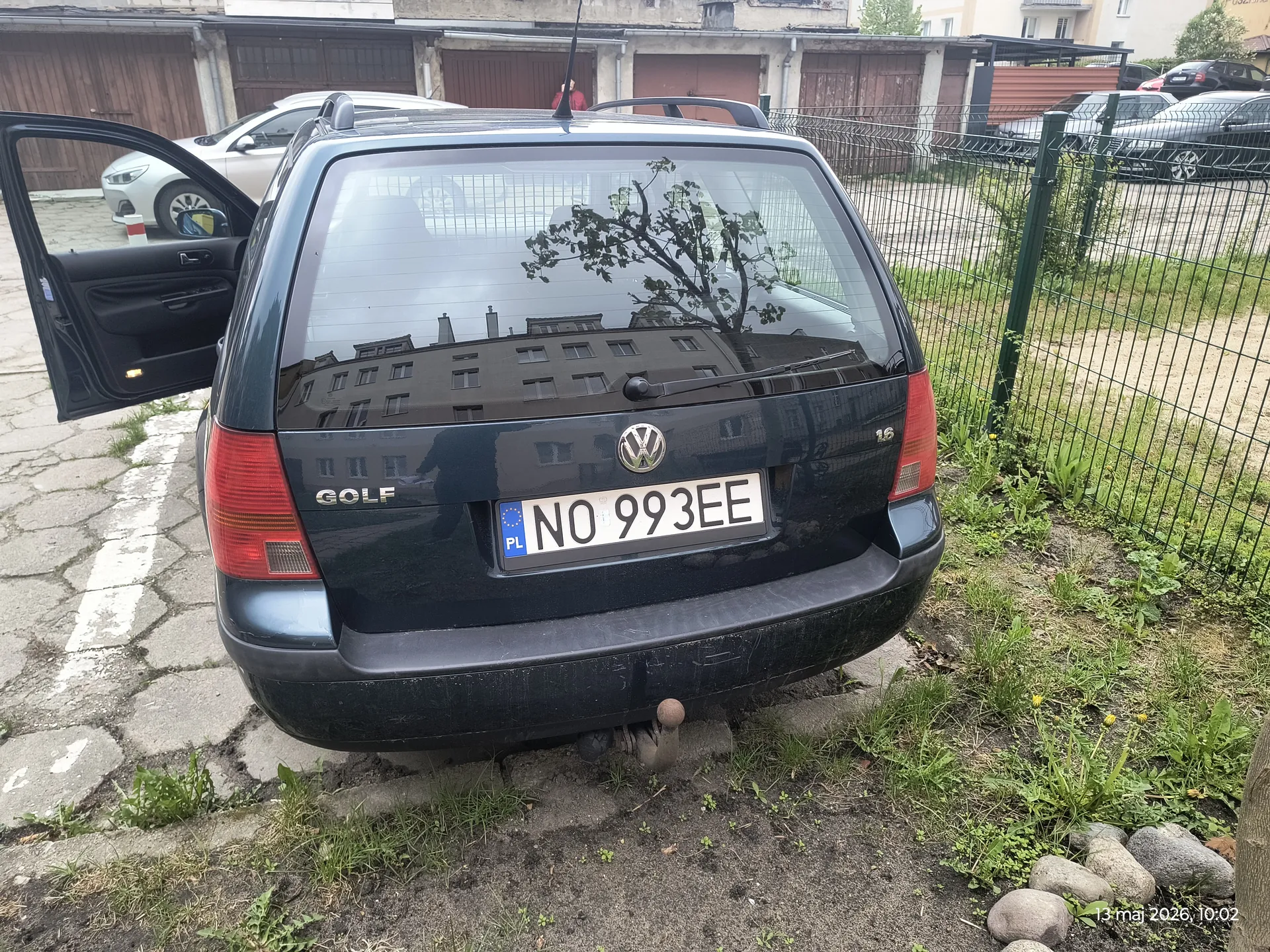 Czarny samochód Volkswagen Golf z tyłu, z otwartą lewą przednią drzwiami. Widoczny tabliczka rejestracyjna PL NO 993EE oraz napis "GOLF" na bagażniku. W tle budynki z brązowymi garażami, siatka ogrodzeniowa, inne zaparkowane pojazdy oraz odbicie budynku w tylnej szybie. Na dole obrazu widoczny znaczek daty i godziny: 13 maj 2026, 10:02. Samochód stoi na brukowanej powierzchni obok trawnika z kamieniami.