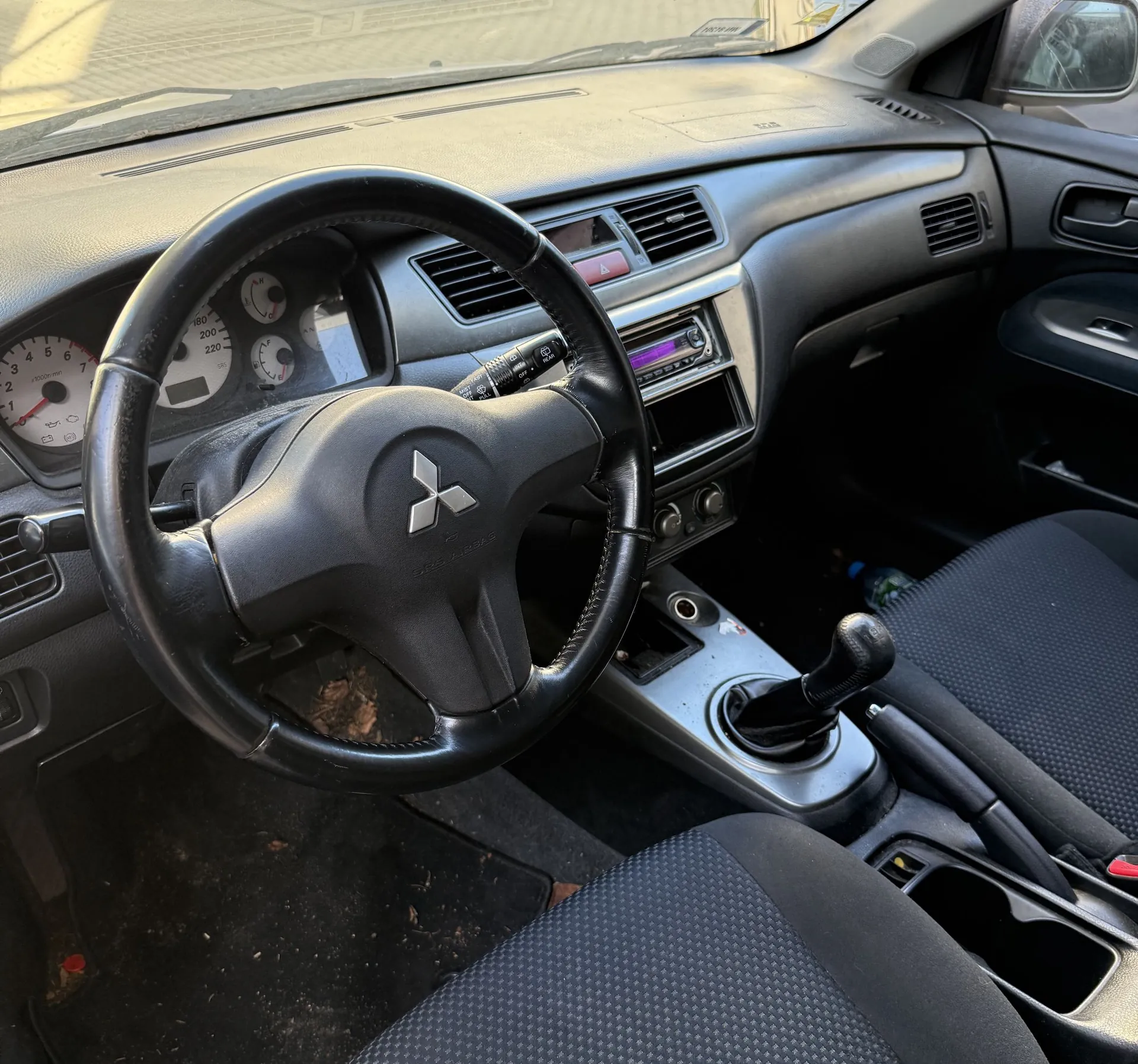 Wnętrze samochodu z perspektywy kierowcy. Widoczna czarna kierownica z logo Mitsubishi, deska rozdzielcza z zegarami, radiem i nawiewami oraz czarne materiałowe fotele. W konsoli środkowej znajduje się drążek zmiany biegów i hamulec ręczny.