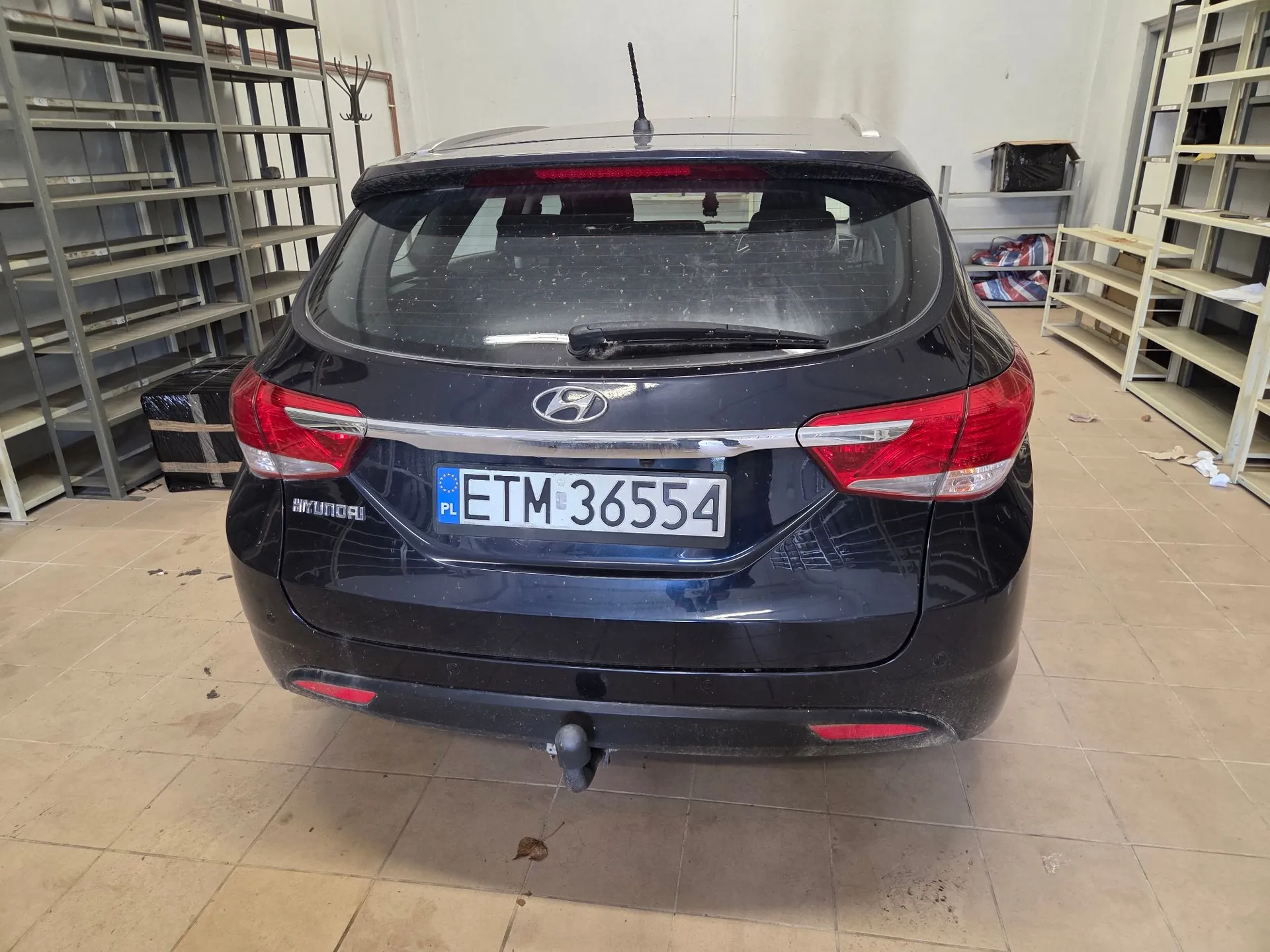 Tylny widok czarnego samochodu marki Hyundai zaparkowanego w pomieszczeniu przypominającym magazyn lub warsztat z metalowymi regałami w tle. Samochód posiada polską tablicę rejestracyjną o numerze ETM 36554 oraz hak holowniczy. Na klapie bagażnika widoczne jest logo Hyundai oraz napis HYUNDAI po lewej stronie.
