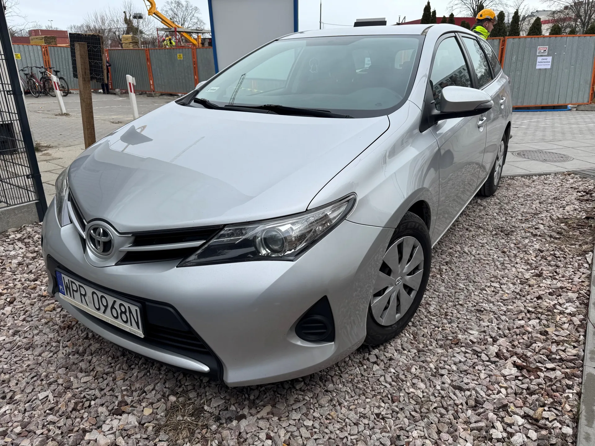 Srebrny samochód marki Toyota z tablicą rejestracyjną WPR 0968N zaparkowany na podłożu z kamieni. W tle widoczne są pomarańczowe ogrodzenia budowlane, żuraw, rowery oraz osoba w żółtym kasku i kamizelce odblaskowej. Na ogrodzeniu umieszczono białą tabliczkę z napisem. Niebo jest zachmurzone.