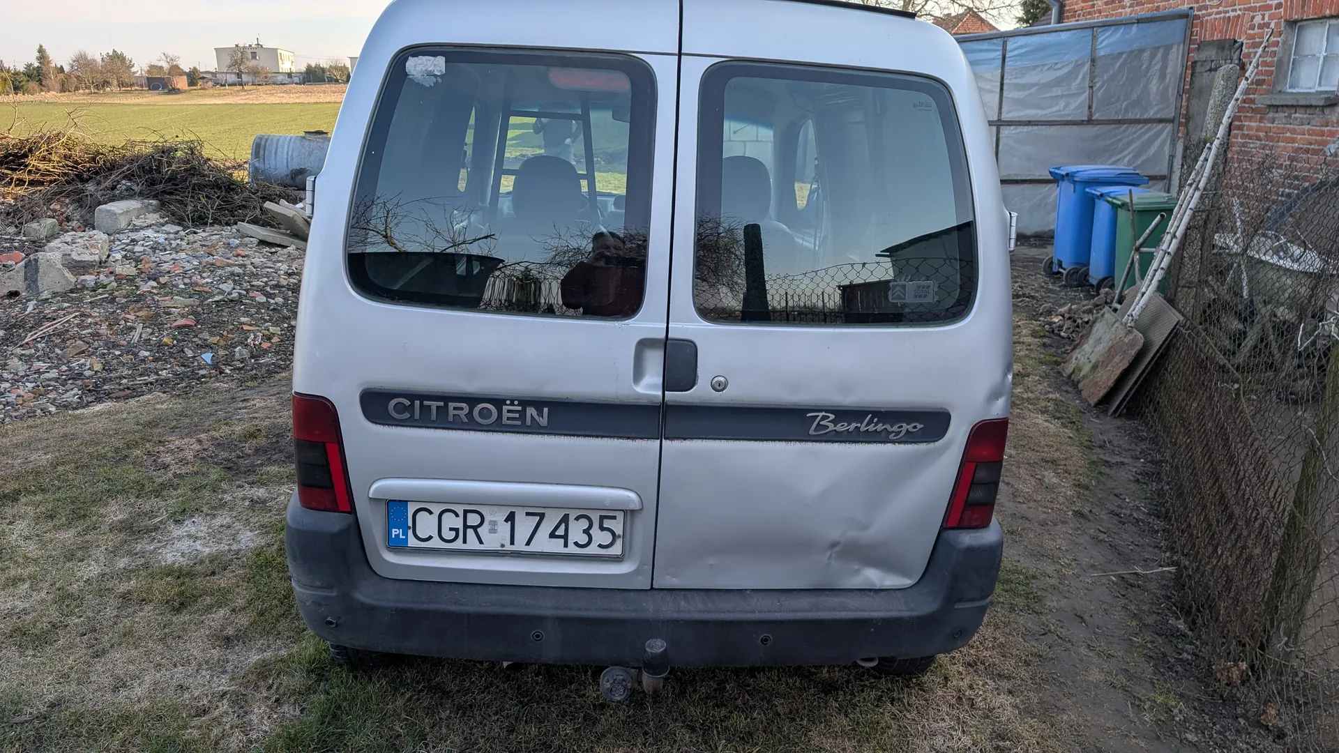 Tył srebrnego samochodu dostawczego Citroën Berlingo z widoczną tablicą rejestracyjną PL CGR 17435. Na tylnej szybie odbicie osoby fotografującej. Po lewej stronie stos gruzu i gałęzi, po prawej niebieskie i zielone pojemniki na śmieci oraz ceglany budynek z siatką. Tło stanowi pole i budynki w oddali. Na tylnym zderzaku widoczny jest wygięty fragment nadwozia.