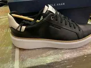 Czarna trampka z białą podeszwą, w której znajduje się drewniana pałeczka. Trampka leży na szklanej powierzchni, w tle niebieskie pudełko z napisem 'SOLE HAAN'.