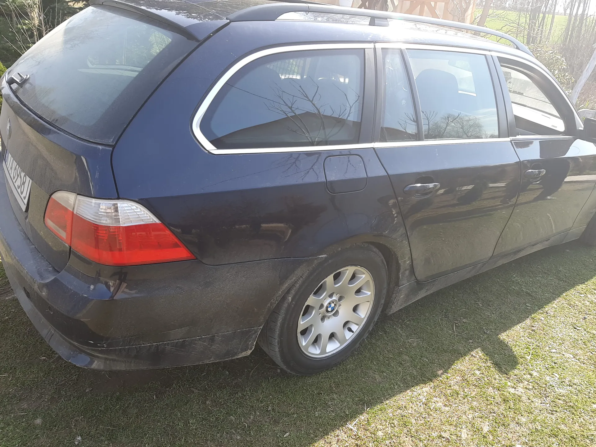 Ciemnoniebieski samochód typu kombi marki BMW, widok z tyłu i lewej strony. Samochód ma zabrudzoną karoserię, czerwone światła tylnego, srebrne felgi z logo BMW oraz tablicę rejestracyjną WA 8958J. Pojazd stoi na trawie, w tle widoczne są drzewa bez liści i fragment budynku. Słońce oświetla scenę, rzucając cień na trawę.