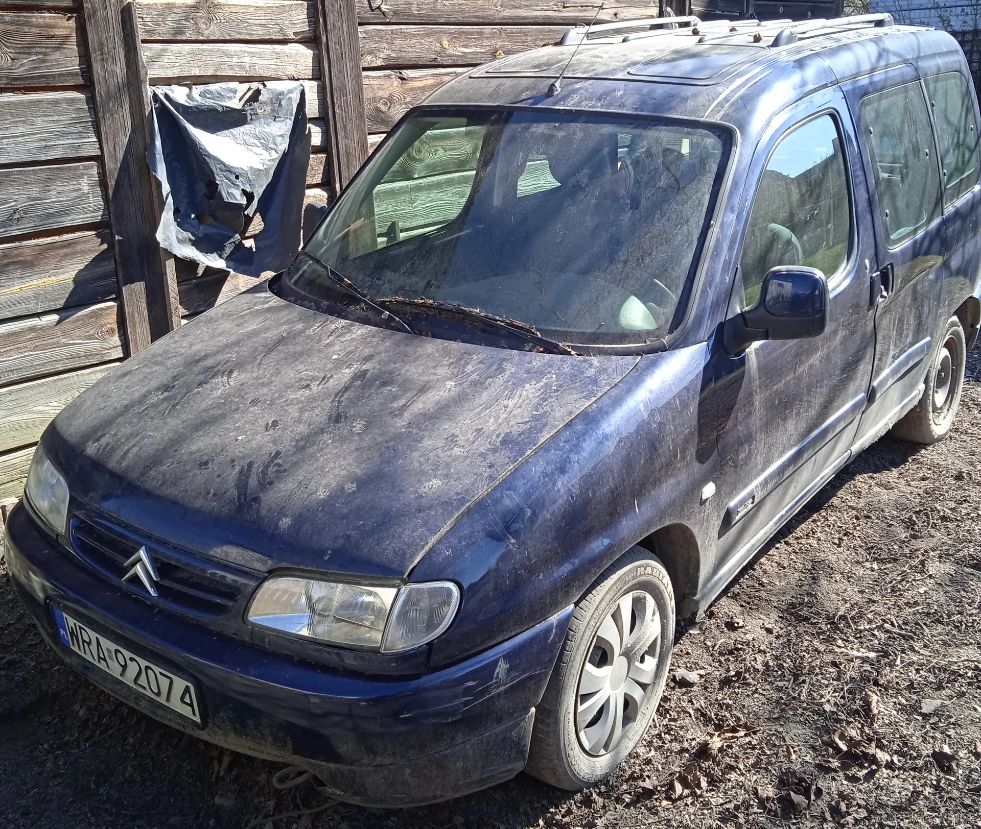 Niebieski samochód osobowy marki Citroën, zaparkowany na podwórku z suchą ziemią i liśćmi. Pojazd jest zabrudzony, na przednim błotniku widoczna tablica rejestracyjna WRA 92074. W tle drewniane ogrodzenie z uszkodzoną folią. Samochód ma dachowy bagażnik i czyste reflektory.